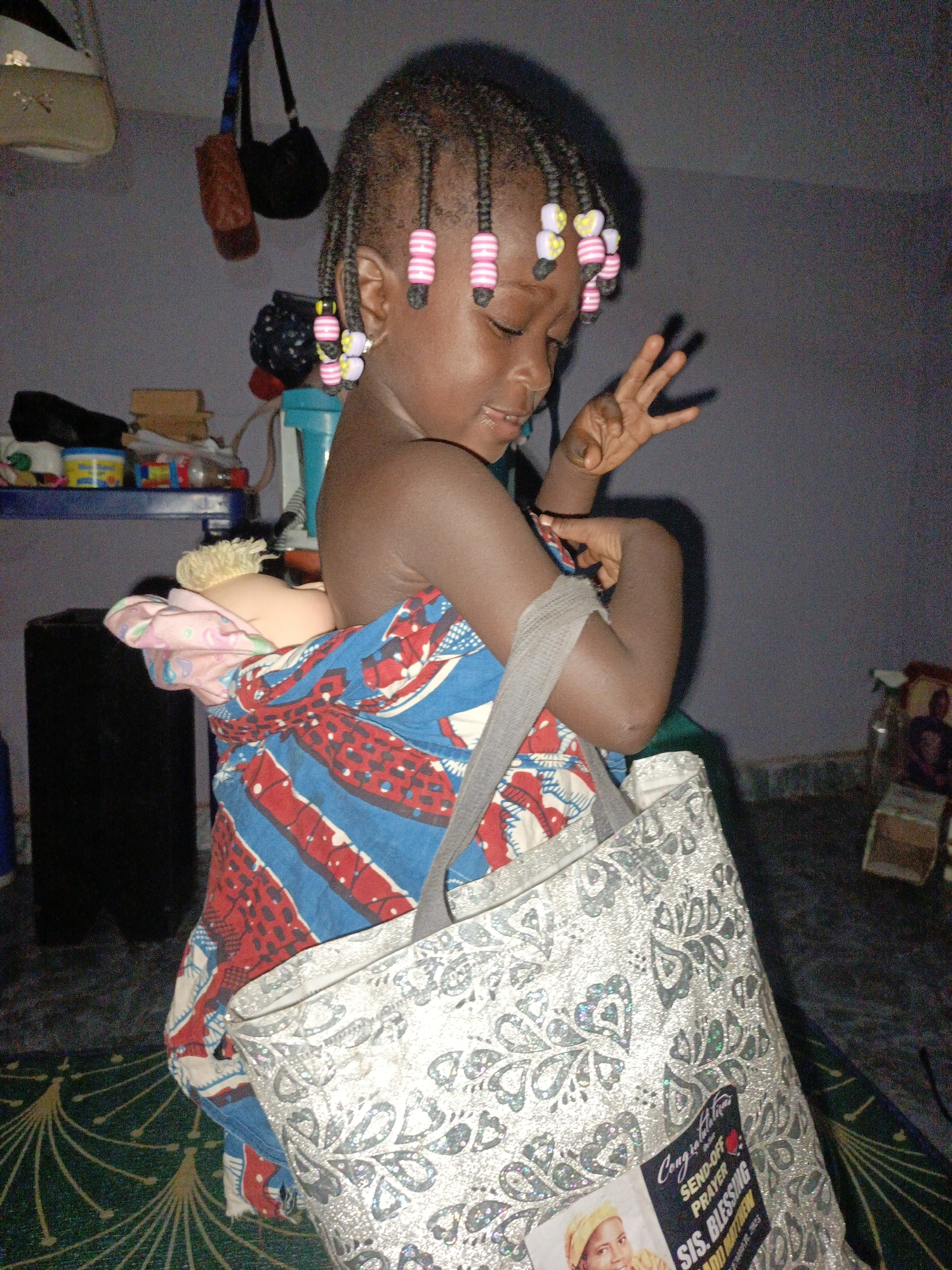 A young Black child, likely female, is captured in a domestic indoor setting. She has dark skin and her braided hair is adorned with numerous pink, white, and yellow beads. The child is focused downwards with a slight smile or thoughtful expression, and her right hand is raised, showing a peace sign with two fingers extended.
She is engaged in playful activity, carrying a light-skinned doll with blonde hair on her back, wrapped in a vibrant blue, red, and white patterned fabric, mimicking the way babies are often carried. The doll's head is visible above her right shoulder. A large, shimmering silver-colored tote bag with an intricate floral or paisley-like pattern is slung over her left shoulder.
The setting appears to be a dimly lit room, possibly suggesting evening or low light conditions. The floor is covered with a green rug or mat featuring a repetitive gold/yellow fan-like pattern. In the background, various household items are visible, including shelves with miscellaneous objects, hanging bags, a black speaker, and a light blue container. A framed picture is also discernible on a small table to the right.
A notable detail is the sticker on the silver tote bag. Visible text on the sticker reads "Congratulations," "SEND-OFF PRAYER," and "SIS. BLESSING." Below this, "ADO MATTHEW" and the date "Saturday 25, 2023" are also clearly legible. The sticker also features a small photo of a person wearing a yellow headscarf. The child's posture and interaction with the doll convey a sense of innocent playfulness.