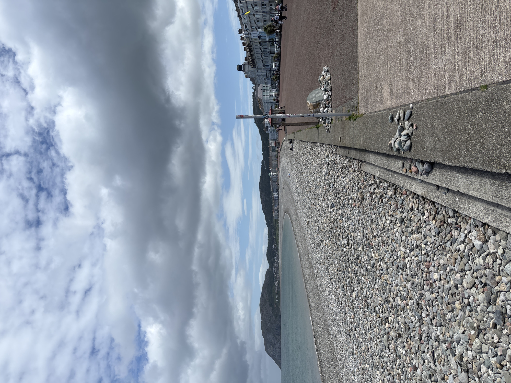 The photograph depicts a pebbled beach with a concrete promenade in Llandudno Junction, United Kingdom. The beach is predominantly covered with grey pebbles. The promenade is constructed with concrete. In the background, there are buildings and a hillside visible under a cloudy sky. The image is oriented vertically.