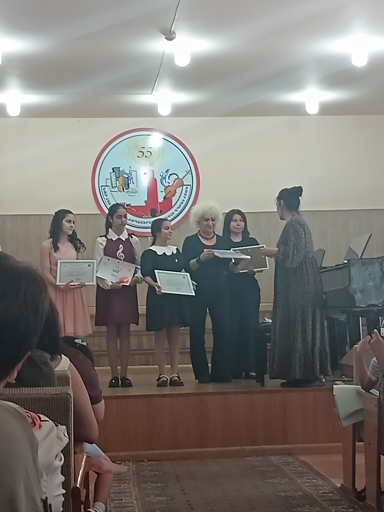 In an indoor setting, a group of people are on a raised wooden platform. A circular emblem featuring the number "55" and musical instruments is visible on the wall behind them. Five women are present on the platform. Three young girls stand on the left, each holding a certificate. The first girl from the left wears a pale pink dress. The second girl wears a dark red dress over a white shirt with a musical note emblem. The third girl wears a dark dress with a white collar. To the right of the girls, two older women in dark clothing are receiving certificates from a woman in a patterned dress who is facing away from the camera. A piano is visible on the far right of the platform. The floor is covered with a patterned rug.