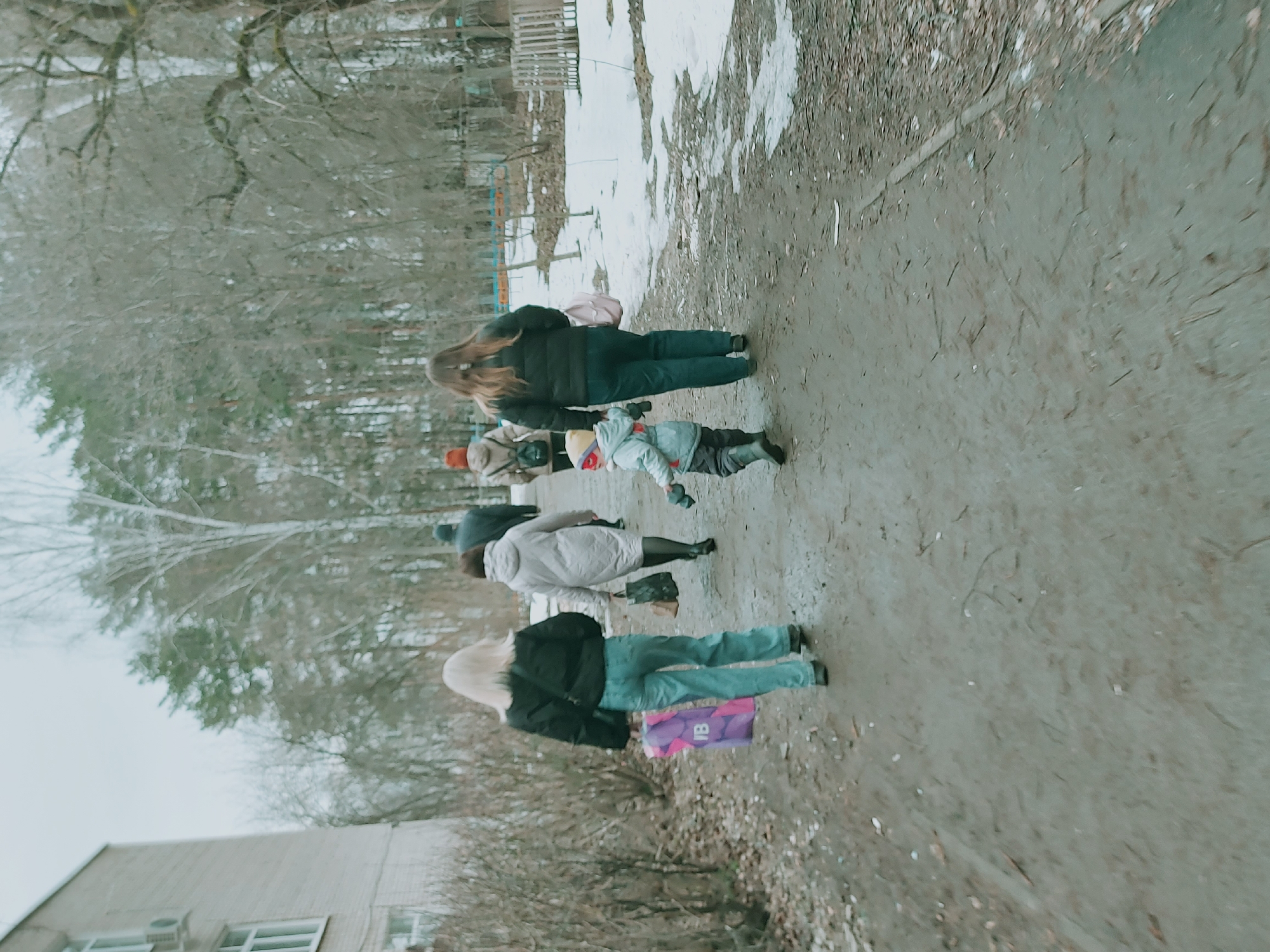 The image shows a group of four people walking along a muddy path in what appears to be a park or outdoor area in Chaikovsky, Russia. The environment suggests a cold climate, with bare trees and patches of snow visible in the background.
The first person in line is a woman with long brown hair, wearing a black jacket and dark pants. Next to her walks a child, bundled in winter clothing. Behind them is a person in a light colored coat, followed by another woman with blond hair, wearing a black jacket and jeans. She is carrying a purple shopping bag. The group is walking away from the viewer, along the path.
To the left of the path is a building with windows. The sky is overcast, contributing to the general impression of a late fall or early spring day. The scene does not convey any specific action other than walking.