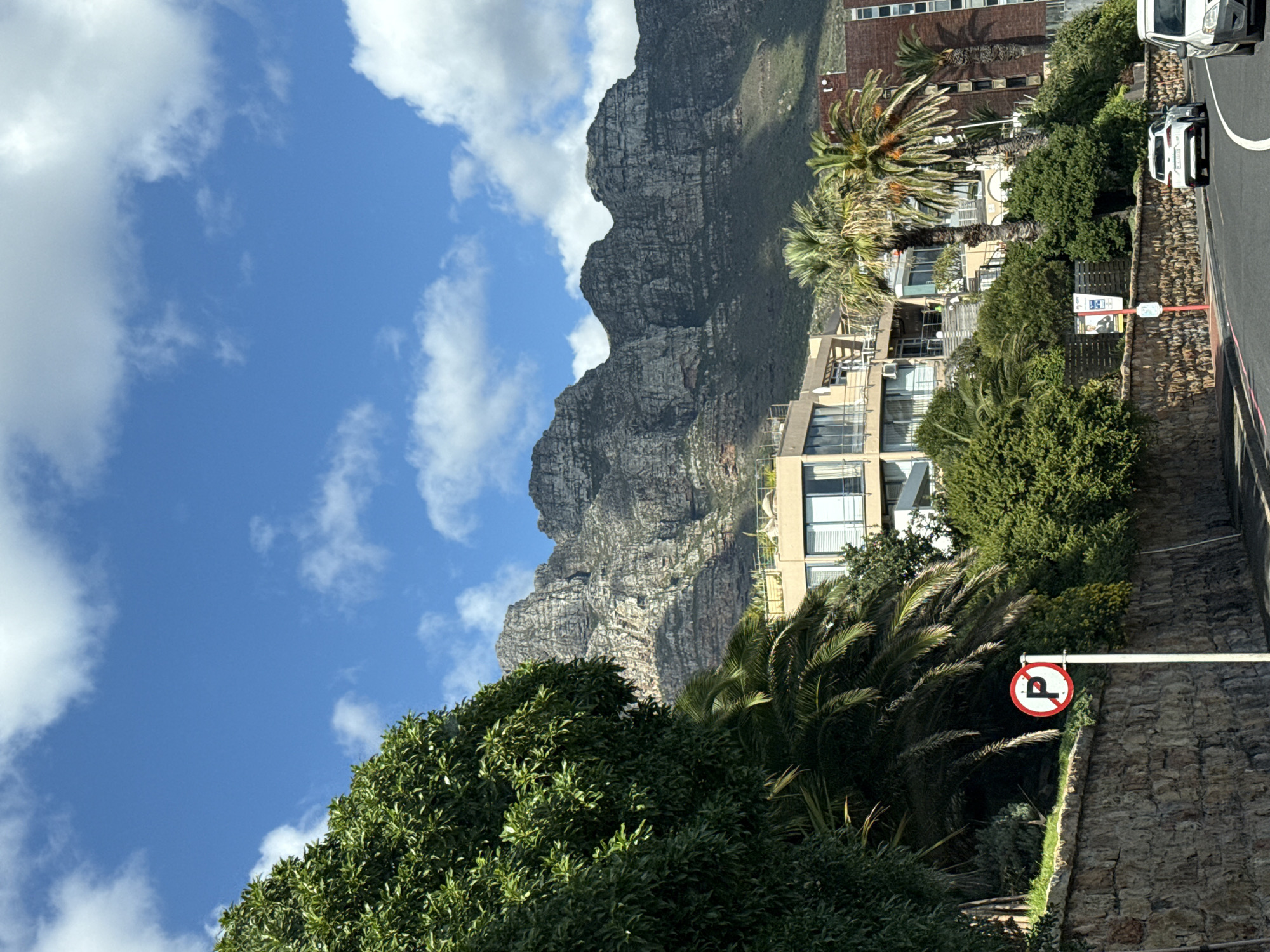 The image presents a vibrant, sunlit urban landscape set against a dramatic natural backdrop, likely in Cape Town, South Africa, given the distinctive mountain profile. The scene is viewed during the daytime with clear weather.
The upper portion of the image is dominated by a bright blue sky dotted with scattered white, fluffy clouds. Below the sky, a massive, rugged mountain range, primarily grey and rocky with patches of green vegetation, rises prominently in the background. Its steep, textured slopes form a majestic natural barrier.
In the mid-ground, nestled at the base of the mountain, are several multi-story buildings, appearing to be modern residential or hotel structures with balconies and large windows. Lush, dense green foliage, including tall palm trees and various bushes, surrounds and partially obscures these buildings, creating a verdant environment.
The foreground on the right features a paved road with a solid white line, running alongside a neatly constructed stone retaining wall. Two white vehicles, an SUV or truck and a sedan, are parked on the side of the road. A white pole extends from the stone wall, holding a circular red and white sign which clearly indicates "No U-turn" with its black arrow symbol. Further along the wall, there is a red, fire hydrant-like object and another white rectangular sign. This sign, when viewed closely, shows a person's face and text that reads "RELAX, WE'VE GOT IT COVERED," suggesting an advertisement.
No direct human activity is visible, presenting a static yet lively scene of urban life integrated with natural beauty. The bright sunlight casts clear shadows, indicating a sunny day.