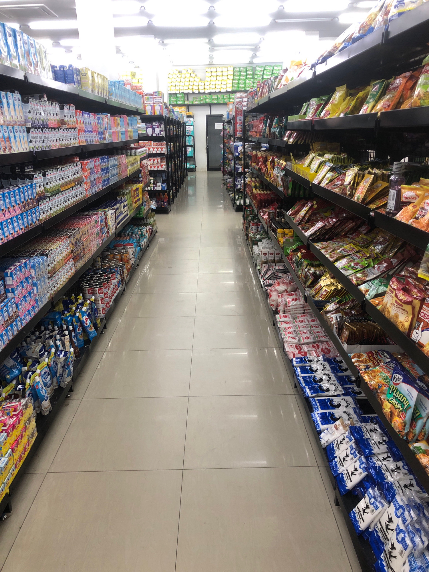 The image shows the interior of a supermarket aisle, likely in Lhokseumawe, Indonesia, filled with shelves stocked with various food products and beverages. On the left side, tall shelves display rows of cartons of milk and juice, along with other packaged drinks and canned goods. On the right side, shelves are packed with an assortment of packaged food items, including snacks, spices, and instant meal kits, many featuring colorful packaging with text in what appears to be Bahasa Indonesia. The aisle is well-lit by overhead fluorescent lights, and the floor is tiled with light-colored square tiles. There are no people visible in the image, and the scene depicts a typical retail environment.