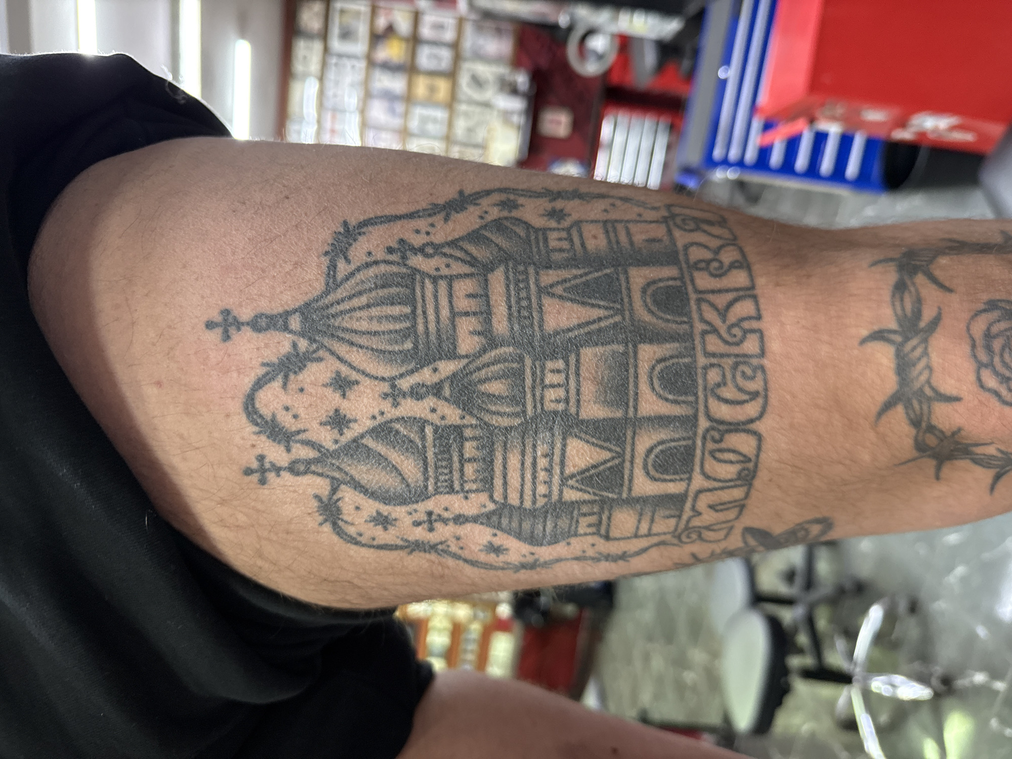The image presents a close-up view of a person's upper arm, featuring a prominent black ink tattoo. The skin tone is light to medium, with some fine hair visible on the arm.
The central tattoo design depicts a stylized rendition of St. Basil's Cathedral, an iconic landmark in Moscow, Russia. The cathedral's distinctive onion domes and intricate architectural details are rendered with shading and line work. Above and below the main structure, the tattoo incorporates elements resembling barbed wire, framing the scene. Within the design, small stars and crosses are visible, adding to the visual texture. To the right side of the cathedral, the word "МОСКВА" (MOSKVA), meaning Moscow, is tattooed vertically in block letters, clearly identifying the city. Further down the arm, a second tattoo of barbed wire wrapping around what appears to be a rose is partially visible.
The background is heavily blurred but suggests an indoor setting, possibly a tattoo parlor or workshop. Faint details include what looks like shelves with framed items on a wall, parts of a red and blue toolbox or cabinet, and metallic elements from what might be a chair or workstation. Bright lights are also visible in the background, creating reflections.
The image captures the tattoo with clear detail, highlighting the artistry and its cultural significance tied to Moscow. No specific activity is shown beyond the arm being photographed.