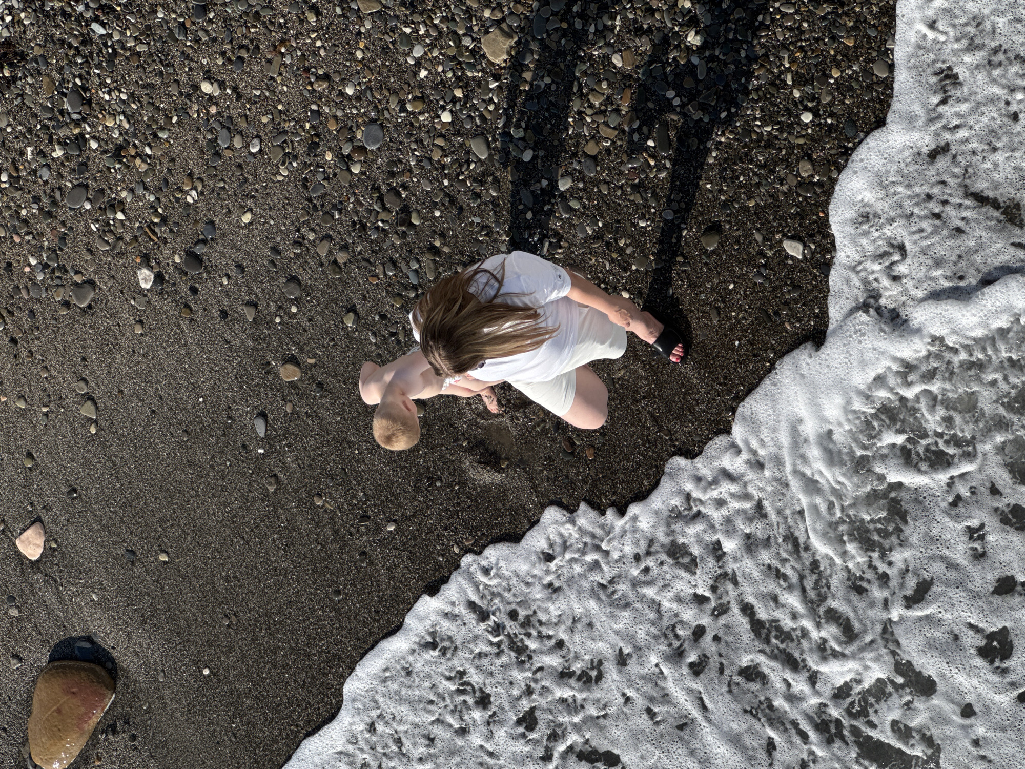 The aerial shot captures a sunny day at a pebble beach in Sochi, Russia. A woman in a white t-shirt and white shorts, wearing black sandals with red nail polish, stands on the dark pebble shore. Beside her, a young child, possibly a toddler, is crouched down, looking towards the water. The sea foam is washing ashore, creating a white foamy line across the dark pebbles and wet sand. In the upper left corner, a large, smooth, reddish-brown stone rests on the dark sand, partially submerged by the incoming water. The scene is one of peaceful beach activity, with the natural textures of pebbles and the dynamic element of the waves.