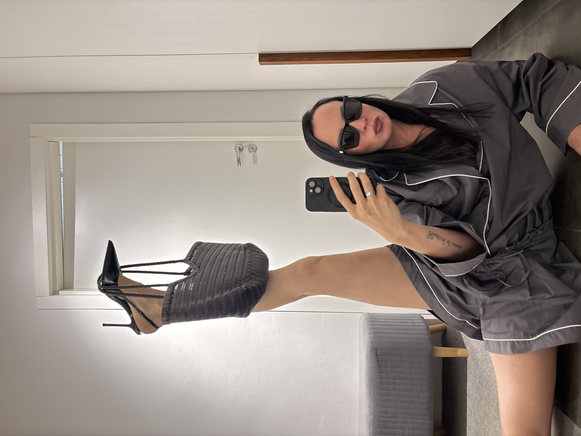 A person is taking a selfie in what appears to be a dressing room or bathroom, with a light-colored door and wall visible. The person has dark hair, is wearing sunglasses, and is holding a black phone. They are wearing a gray pajama-like set with white piping. The individual's right leg is extended upwards, adorned with a black stiletto heel and a black woven handbag. A gray padded stool is located near the leg. Based on the visual details, no information suggests the location is Chaikovsky, Russia.