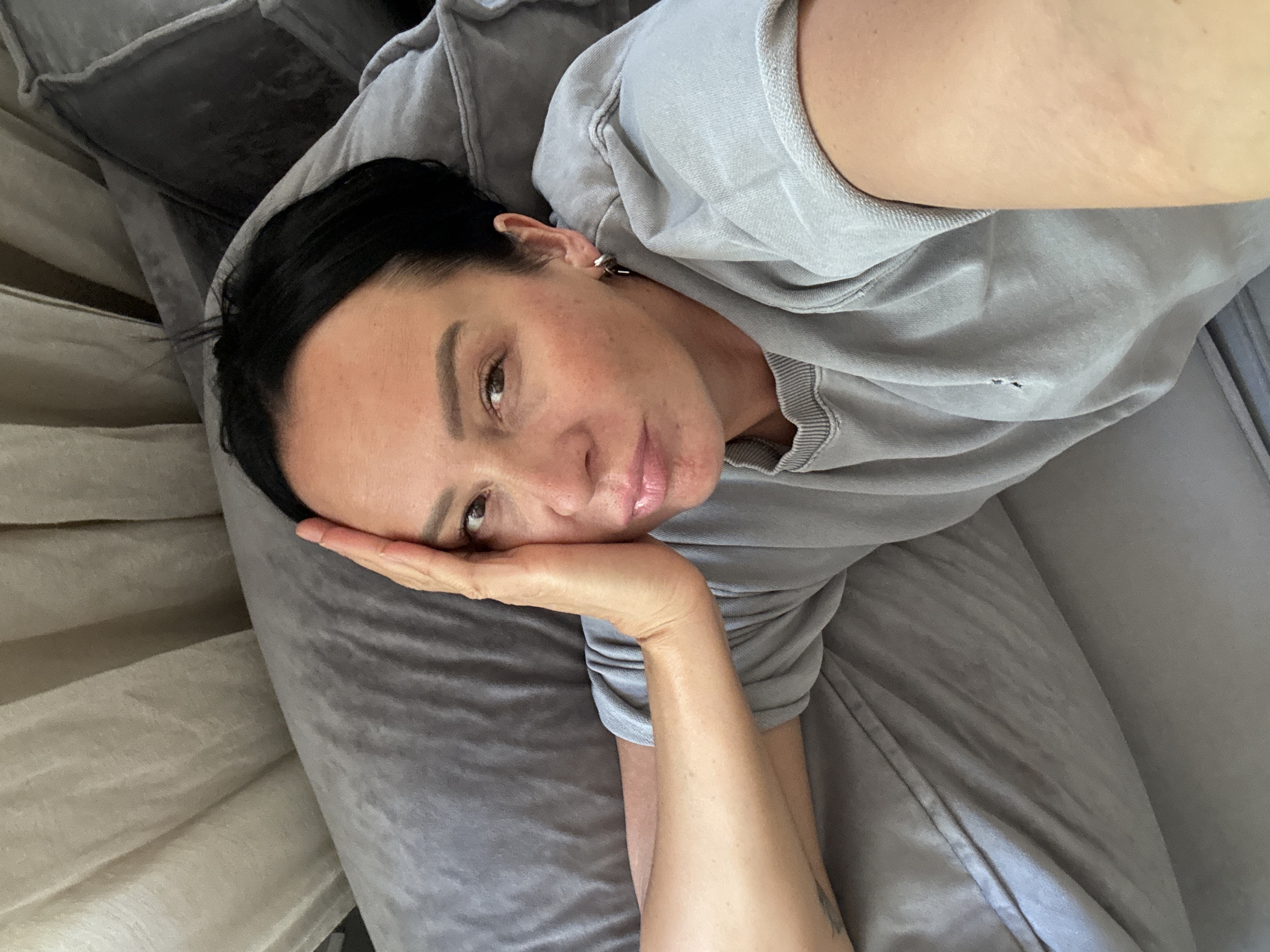 This media file is a portrait of a person lying on their side, propping their head up with their hand. The person has dark hair and is wearing a grey sweatshirt. They are looking at the camera with a relaxed expression. The background appears to be a bed with grey pillows. The lighting suggests it is daytime. There is no visible text in the image.