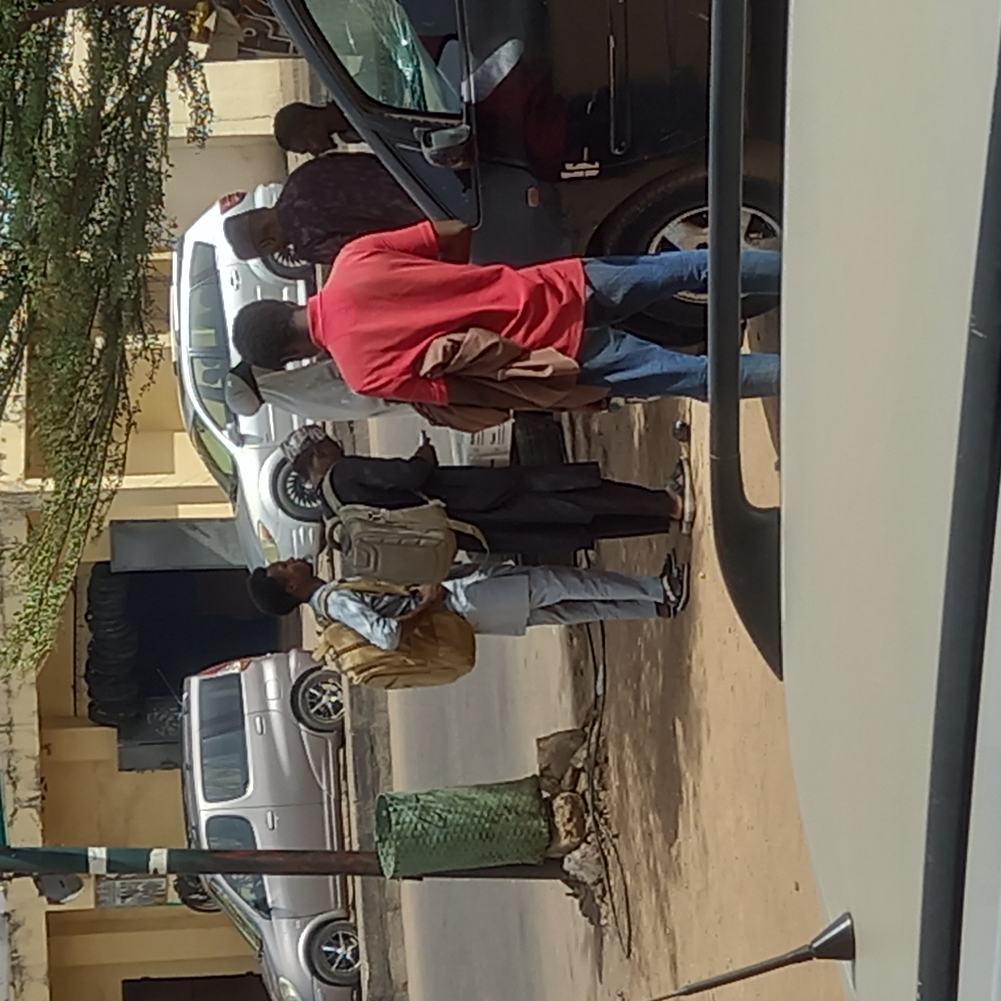 This outdoor image, taken in Dadi Riba, Nigeria, depicts several men gathered around parked cars on a sunny day. The scene appears to be a street or roadside with dusty ground and light-colored buildings in the background.
In the immediate foreground, a man in a red short-sleeved shirt and blue jeans is leaning into the open front door of a dark-colored car, possibly an SUV. A brown garment is draped over his shoulder. To his left, another man in dark clothing wears a light-colored backpack and is looking towards the car. Next to him, a third man, dressed in a light blue top and light-colored trousers, also carries a light-colored backpack and looks upwards. A fourth man's head is partially visible behind the dark car's open door, and another person can be seen further back behind a white car.
Beyond the dark car, a white sedan is visible, and further down the street on the left, a silver compact SUV or minivan is parked. The left side of the frame features a tree with dense green foliage. Along the dusty ground, there's a green woven, cylindrical object situated near a dark pole, surrounded by some rocks or debris. The bright sunlight indicates it is daytime, likely mid-morning or afternoon, with clear weather conditions. No discernible text is visible. The activity appears to be a casual stop or interaction beside the cars.