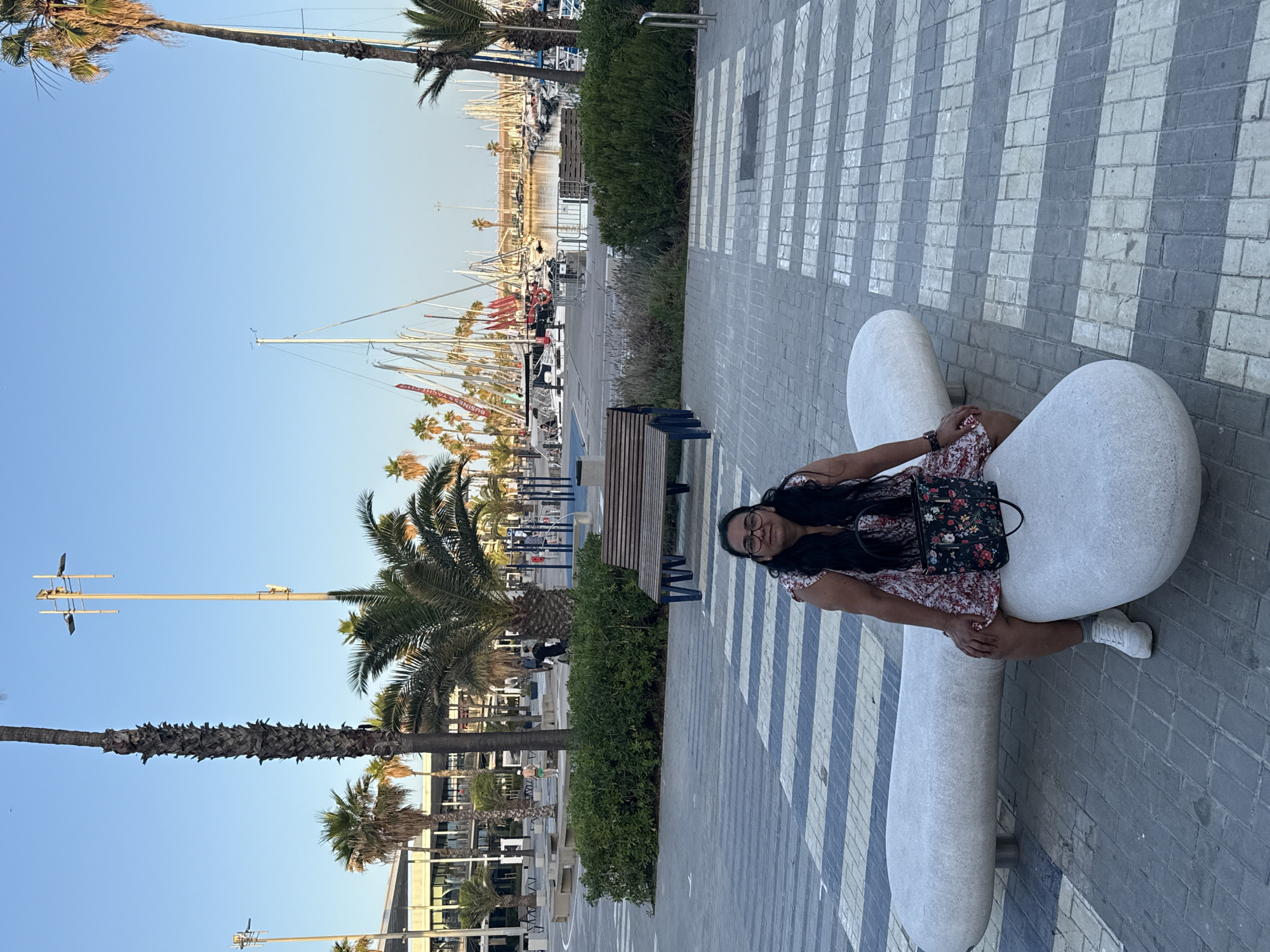 A woman sits on a white, elongated bench in front of a paved walkway with white stripes in a modern, open urban area. She is wearing a floral print dress, glasses, and white sneakers, with a black handbag on her lap. The bench is positioned near a large planter filled with green bushes and a wooden bench. In the background, palm trees and tall light poles are visible. Beyond the trees, a harbor filled with sailboats and yachts stretches out under a clear blue sky, with a modern building complex to the left. The scene suggests a waterfront location, likely a marina or promenade.