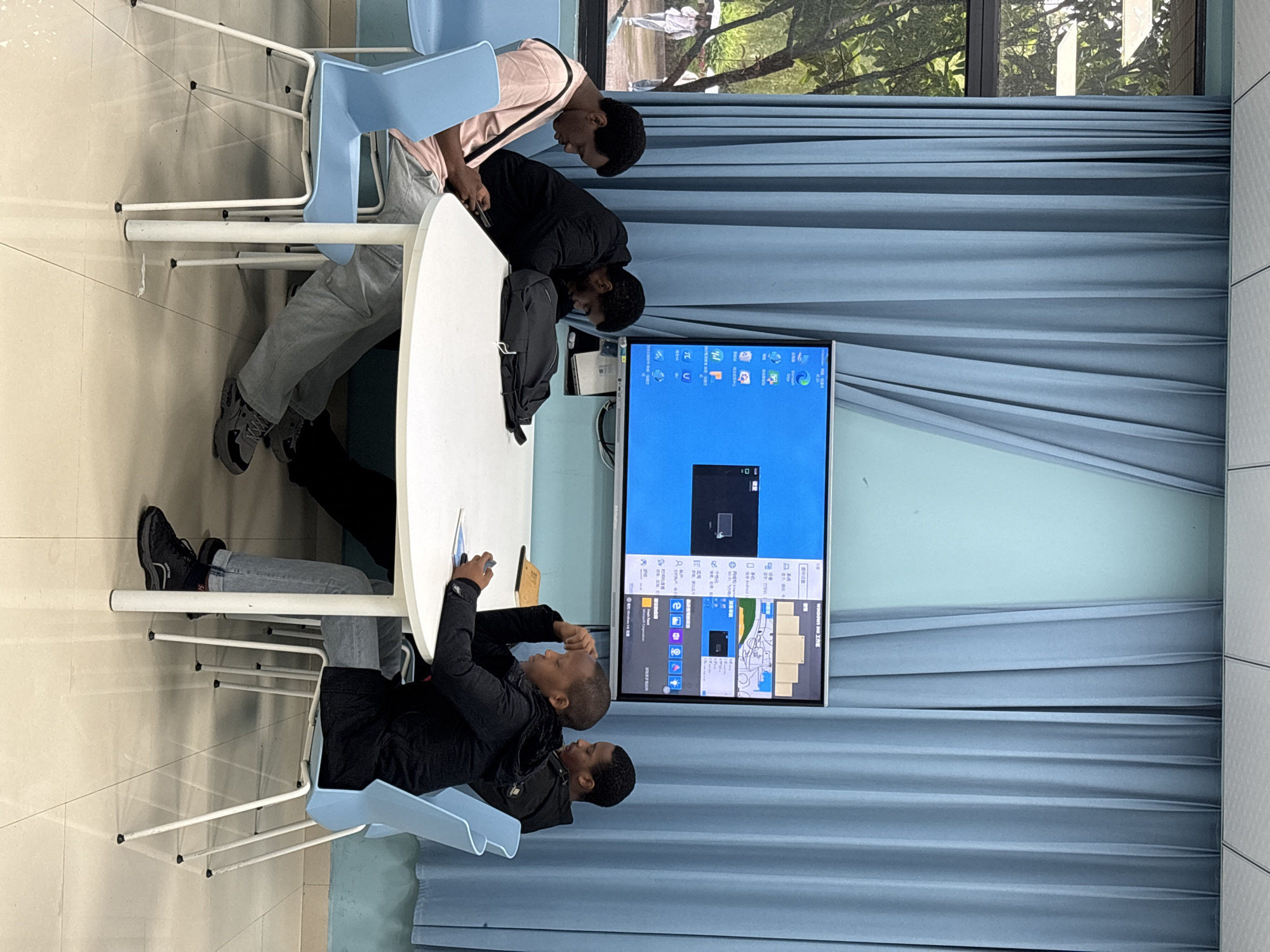 In a tiled room, four people are gathered around an oval, white table. They are examining a screen showing a computer desktop interface. The people are seated on light blue chairs with white legs. A window behind the screen provides a view of trees. Blue curtains cover part of the wall behind the screen. The location is Jiangjin, China.