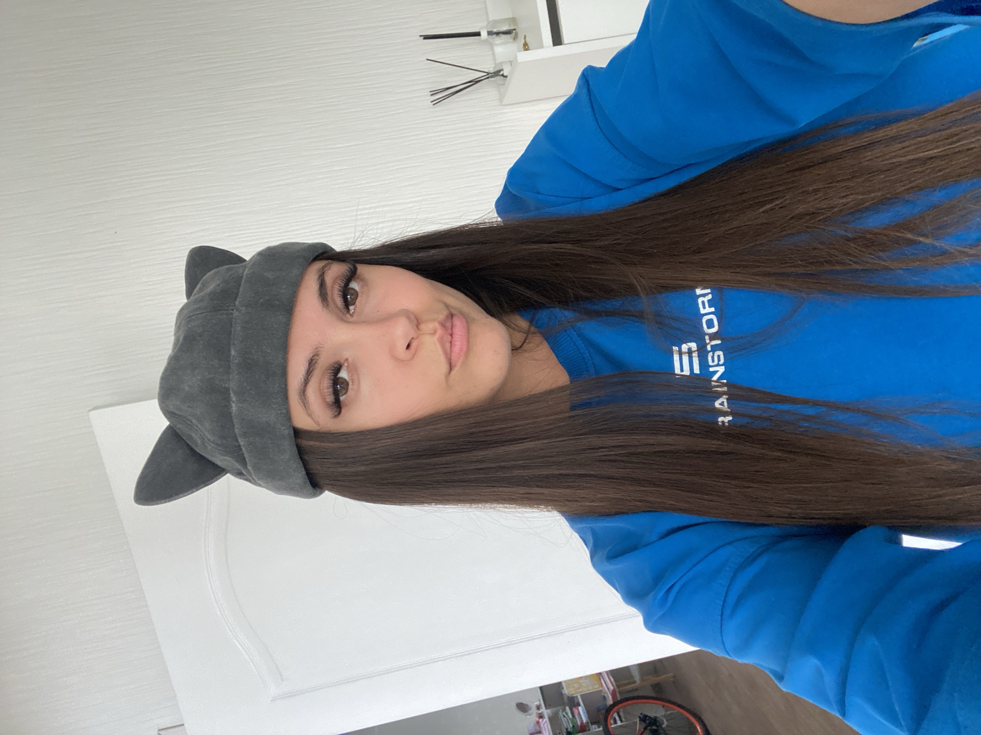 A young woman with long, dark hair and prominent eyelashes is shown from the chest up, looking directly at the camera. She is wearing a gray hat with cat-like ears and a blue sweatshirt with white lettering on the left side of her chest that reads "RAINSTORM". Her facial expression is neutral with slightly pursed lips. The background appears to be an indoor setting, possibly a home, with a white wall and door visible. A shelf with small objects is partially visible in the background on the right. The lighting suggests it is daytime.