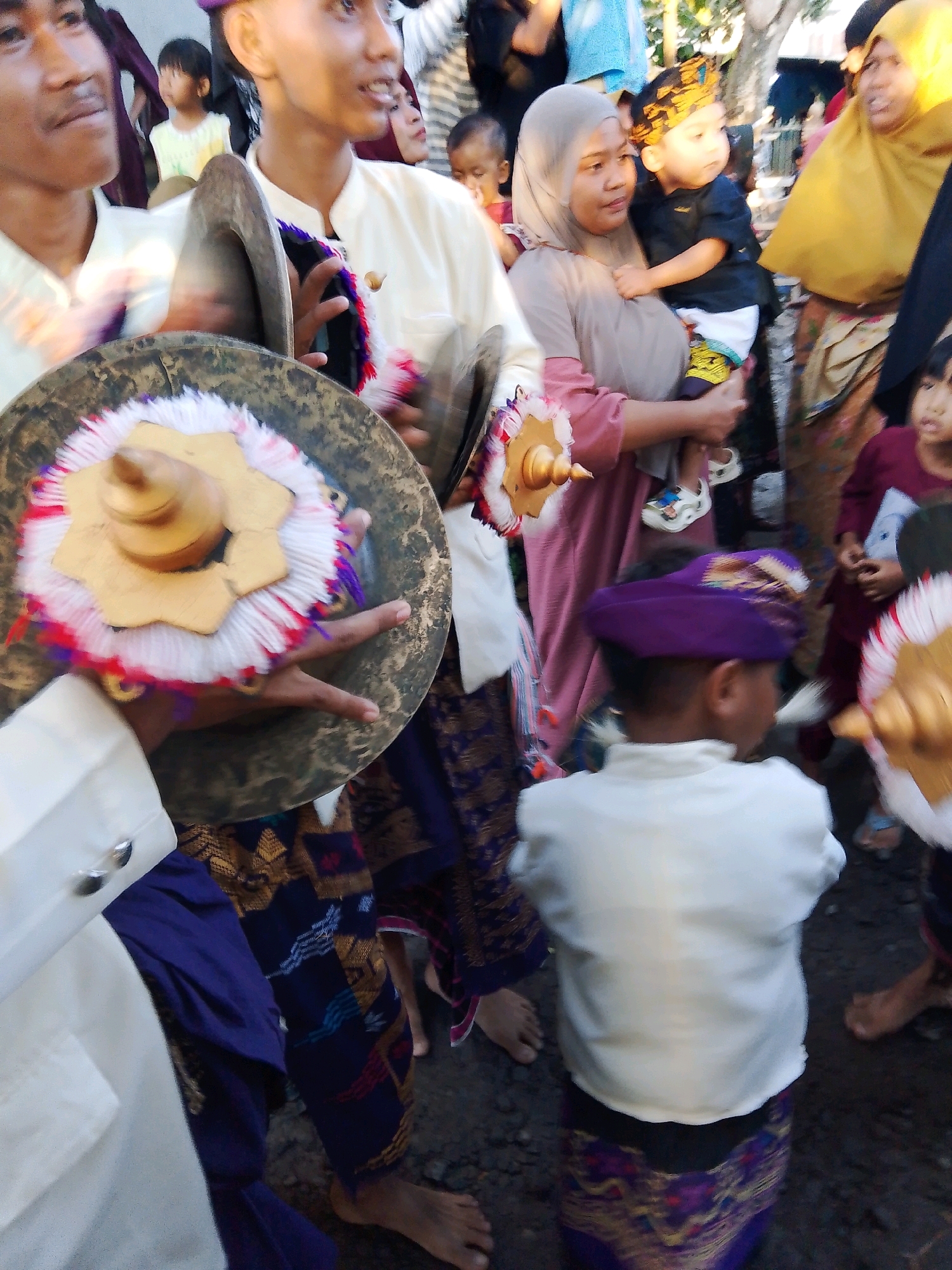 The image depicts a cultural event, likely a procession or celebration, in Kuripan Utara, Indonesia. In the foreground, a person in a white shirt and purple patterned sarong is holding a gong adorned with white, red, and purple tassels. To the left, another individual in a white shirt is also holding a gong. In the middle ground, a woman wearing a hijab holds a young child dressed in a dark shirt and patterned pants. Other individuals, including children and adults, are visible in the background, some wearing traditional attire. The setting appears to be an outdoor, possibly crowded, area with natural light suggesting it's daytime. The ground is uneven and appears to be dirt or gravel.
