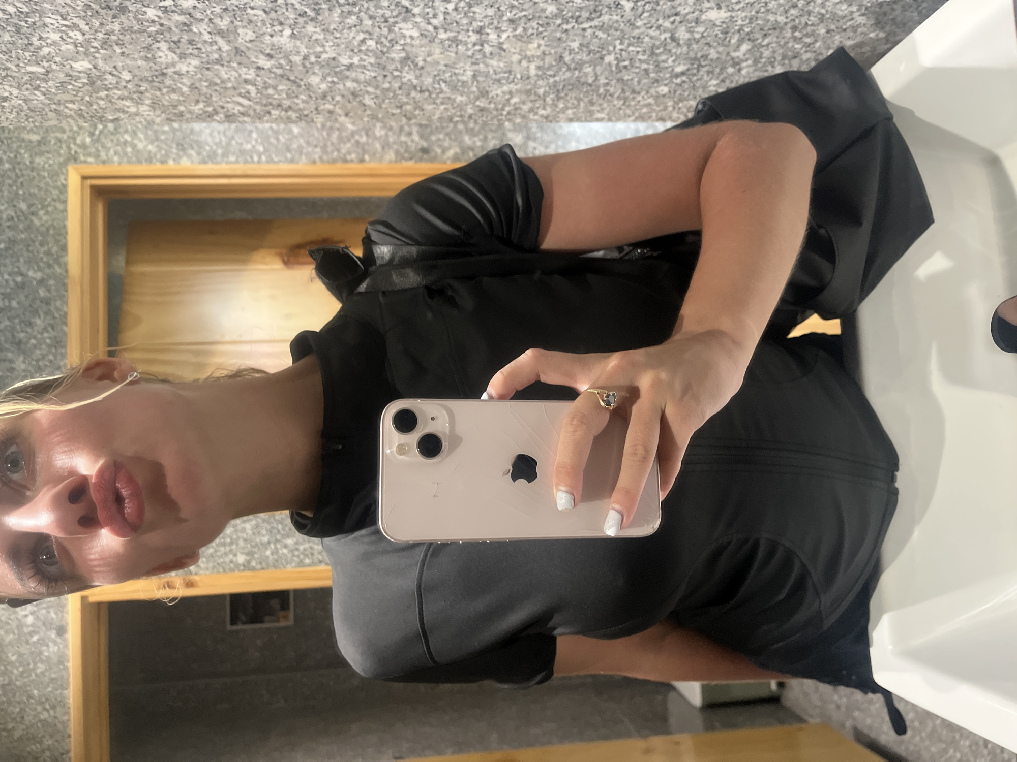 A woman is taking a selfie in a bathroom. She is wearing a black short-sleeved top and has blonde hair. She is holding an iPhone in her right hand. She has on a ring on her left ring finger. The bathroom has a granite-patterned wall, a wooden door, and a white sink.