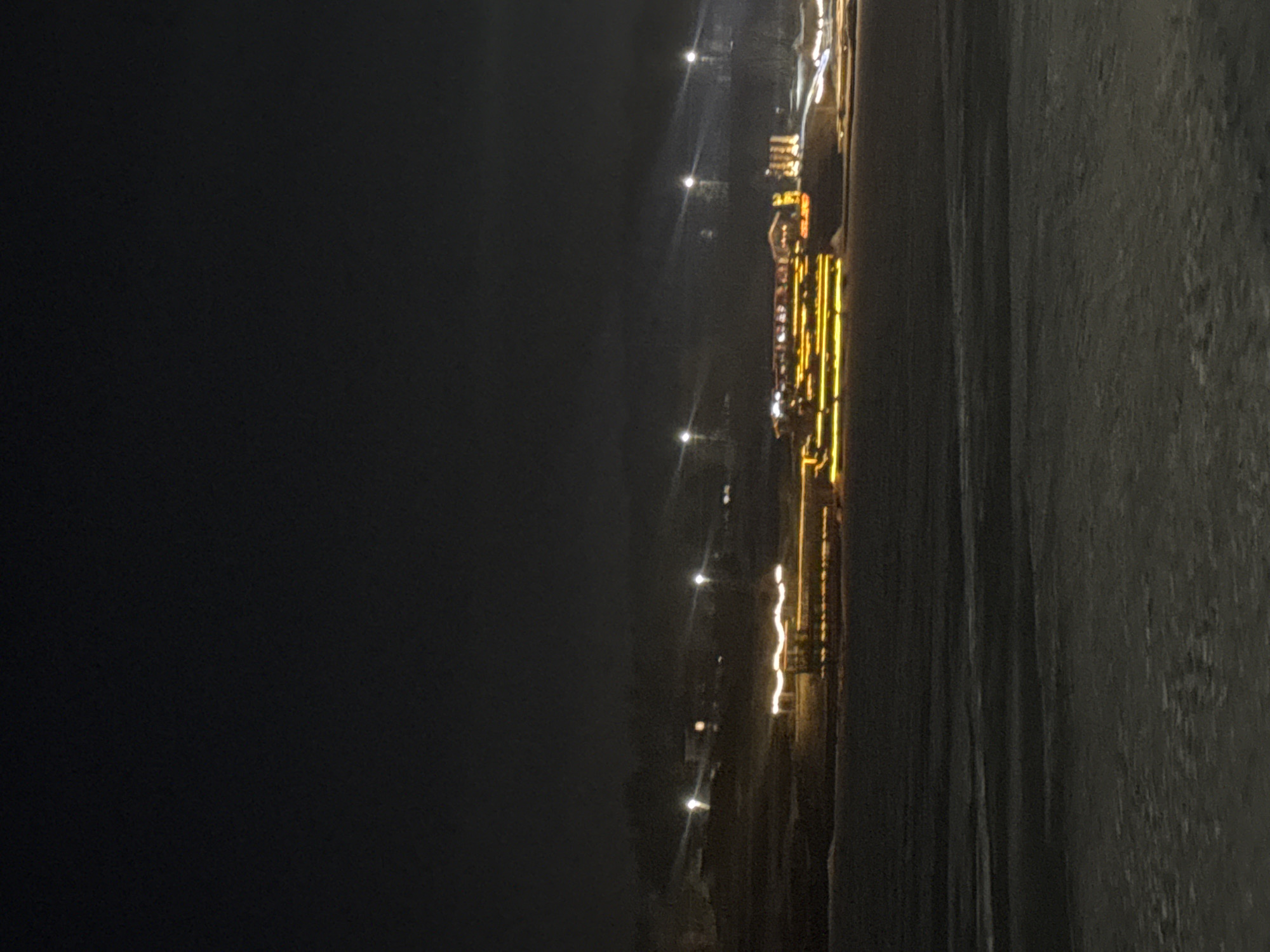 At night, a long pier stretches along the edge of a body of water, possibly a bay or inlet, in Votkinsk, Russia. The pier is lined with numerous bright yellow lights and some illuminated buildings. The area to the left is shrouded in darkness, while the body of water to the right reflects some light, suggesting it may be a calm night. No individuals are visible in the image.