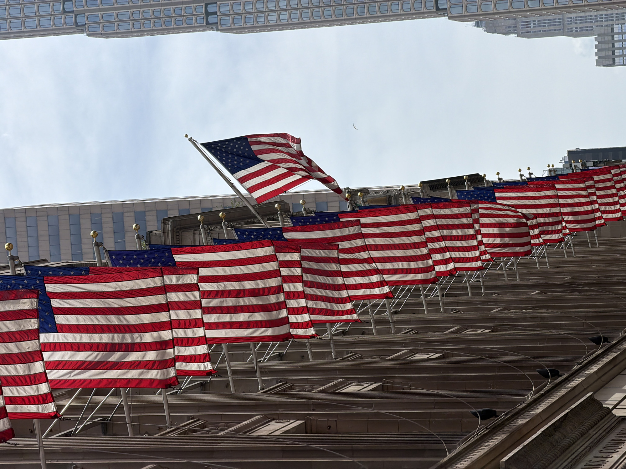 The image depicts a row of American flags displayed on the exterior of buildings in New York City. The flags hang vertically from poles attached to the buildings, creating a patriotic display. The architecture of the buildings suggests a commercial or financial district. In the background, modern skyscrapers rise against a lightly clouded sky. One flag, closer to the foreground, is unfurled and extended outwards, its stars and stripes clearly visible. The scene is likely captured on a daytime, possibly a holiday or commemorative occasion.