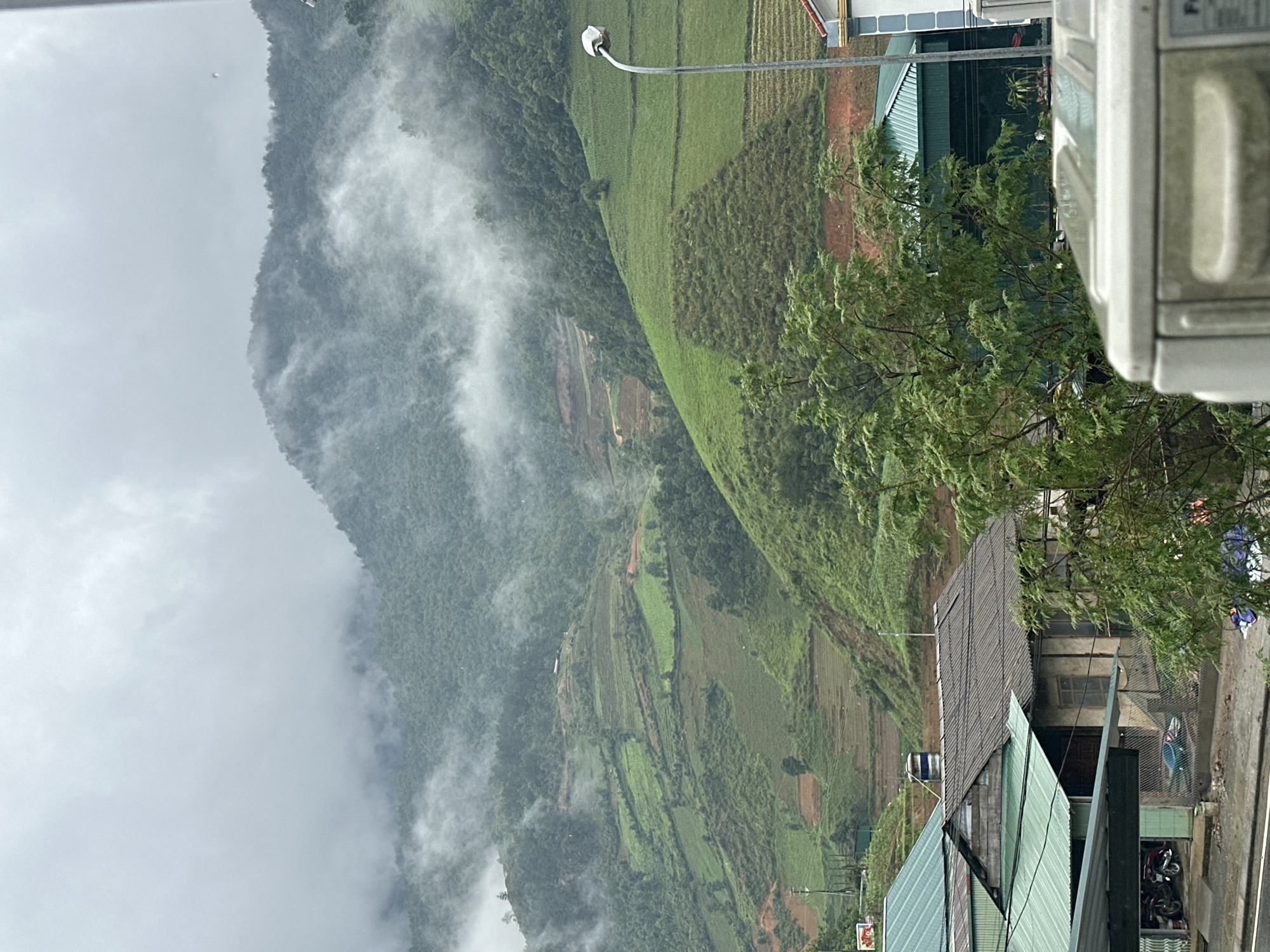 This media file depicts a picturesque mountainous landscape in Mù Cang Chải, Vietnam, blending natural beauty with rural human settlement, under overcast and misty conditions.
In the foreground, to the right of the image, the side of a white outdoor air conditioning unit or similar appliance is partially visible. A prominent green tree with abundant foliage occupies the mid-right, extending over several buildings. Below the tree, a cluster of buildings with distinct corrugated metal roofs, some painted green and others grey or brown, line what appears to be a road or path. A blue water barrel is visible on one roof, and motorcycles are parked near another building. A street light or utility pole extends upwards from the right side of the frame. Blurry human figures are discernible in the distance near the road. Partial text, possibly "SA" or "SI," is visible on a red and white sign, and a green sign is also present.
The midground showcases rolling hills and mountains carved into extensive green agricultural terraces, characteristic of rice paddies or other crops, interspersed with forested areas. Small structures, likely homes or sheds, are nestled among these terraces, and dirt paths snake through the cultivated land.
The background is dominated by steeper, densely forested mountains partially shrouded in thick, low-hanging mist or clouds, which lend a serene and somewhat mysterious atmosphere to the scene. The diffused light suggests an overcast day, possibly early morning or late afternoon. The terraced landscapes confirm the location in Mù Cang Chải, Vietnam.