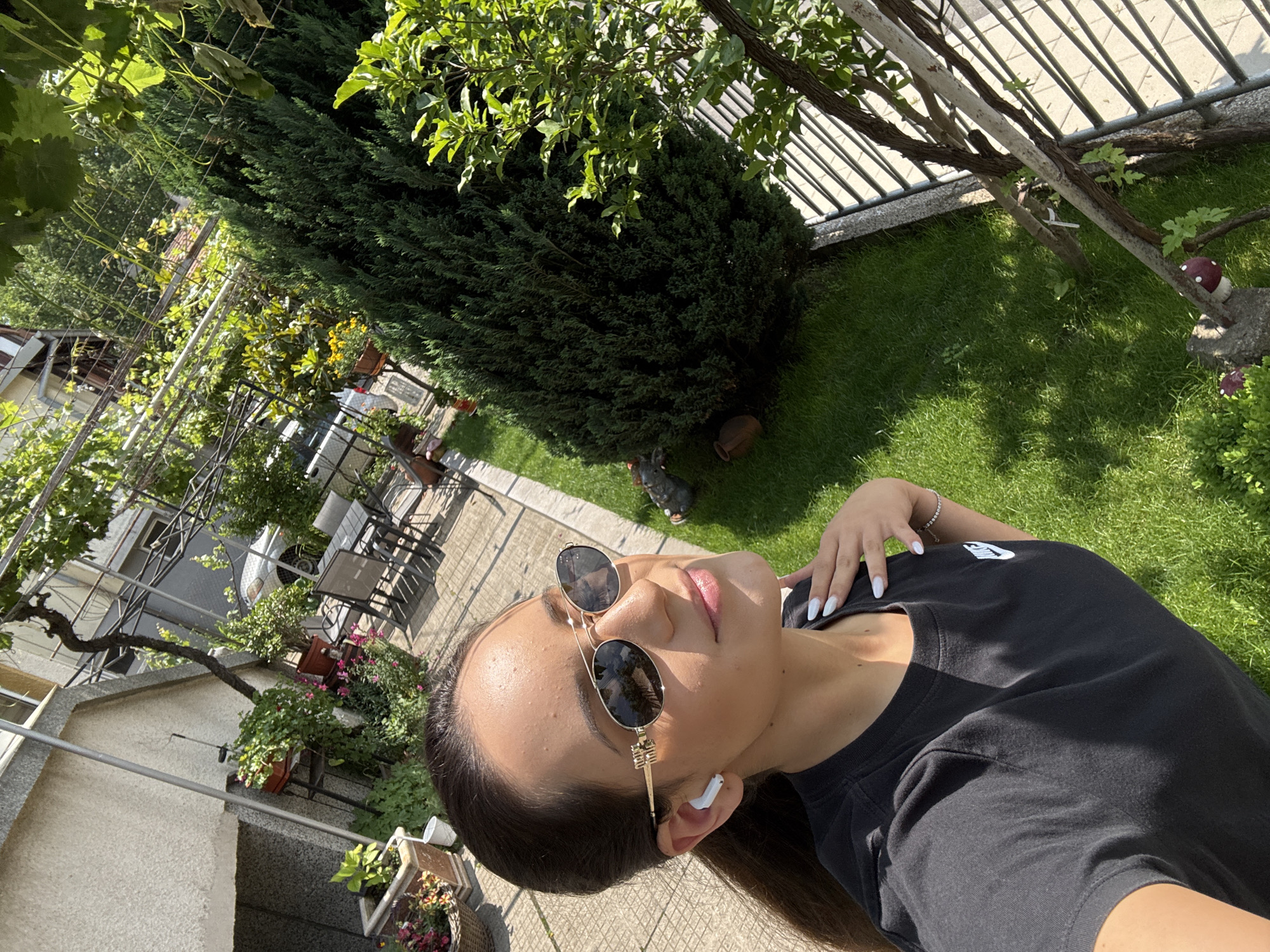 The image displays a bright, sunny outdoor scene, presented from a rotated perspective. The primary subject is a young person, likely female, positioned in the foreground. She has dark hair and is wearing dark, round sunglasses with gold-colored frames, and white wireless earbuds are visible in her ears. Her expression appears relaxed, with her lips slightly parted. She is dressed in a black t-shirt featuring a small white logo, possibly Nike, on the left chest. Her fingernails are painted a light, likely white, color, and she wears a silver-toned bracelet on her right wrist, which rests gently on her chest.
The background reveals a well-maintained garden or yard. Green grass covers a significant portion of the ground. A dense, tall dark green conifer or hedge is prominent in the mid-ground, with a lighter green deciduous tree or vine behind it on the left. A metal fence is visible further back. To the left, parts of a light-colored building with windows and an outdoor seating area with tables and chairs are visible, partially obscured by lush green foliage and what appears to be a pergola structure. Potted plants containing colorful flowers are also present. A rustic brown ceramic pot sits at the base of the large hedge. On the right, nestled in the grass, a decorative red and white spotted mushroom sculpture rests on a small rock, with another smaller, dark red object nearby. The clear, bright sunlight indicates it is daytime. The scene suggests a relaxed moment, possibly a selfie, enjoying a pleasant outdoor environment. No other discernible text is visible.