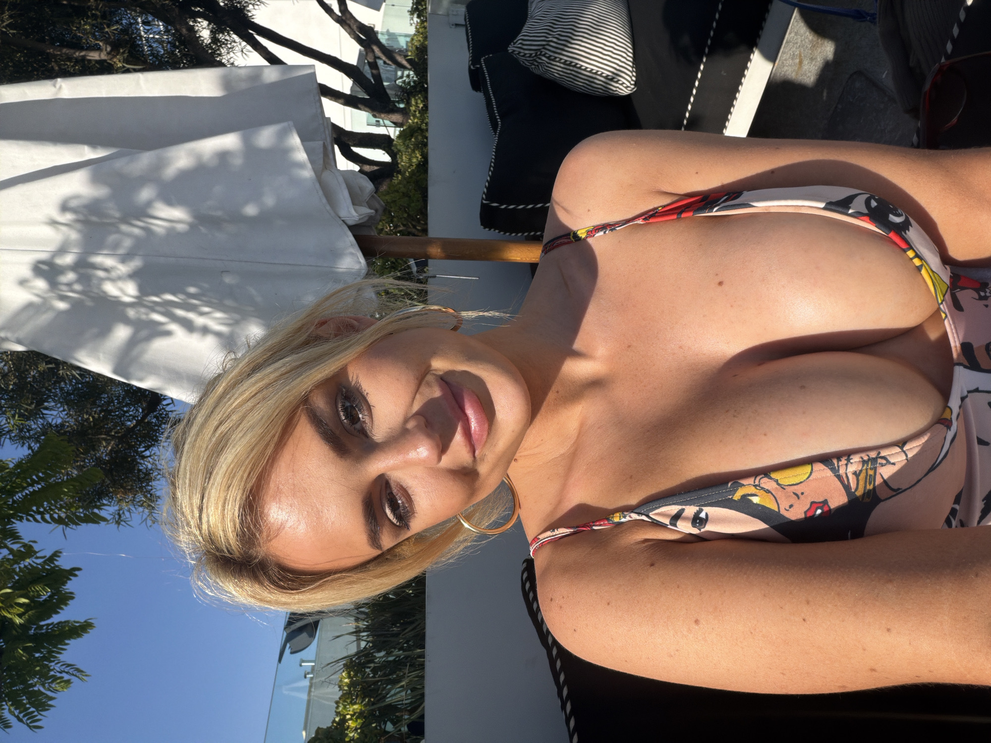 A blonde-haired woman is posing in a swimsuit outdoors. The woman is wearing a swimsuit. She has blonde hair and is wearing gold hoop earrings. She is looking at the camera and smiling. The background includes a white umbrella, some trees, and a building. The image was most likely taken in Los Angeles, United States of America, because of the architectural style of the building.