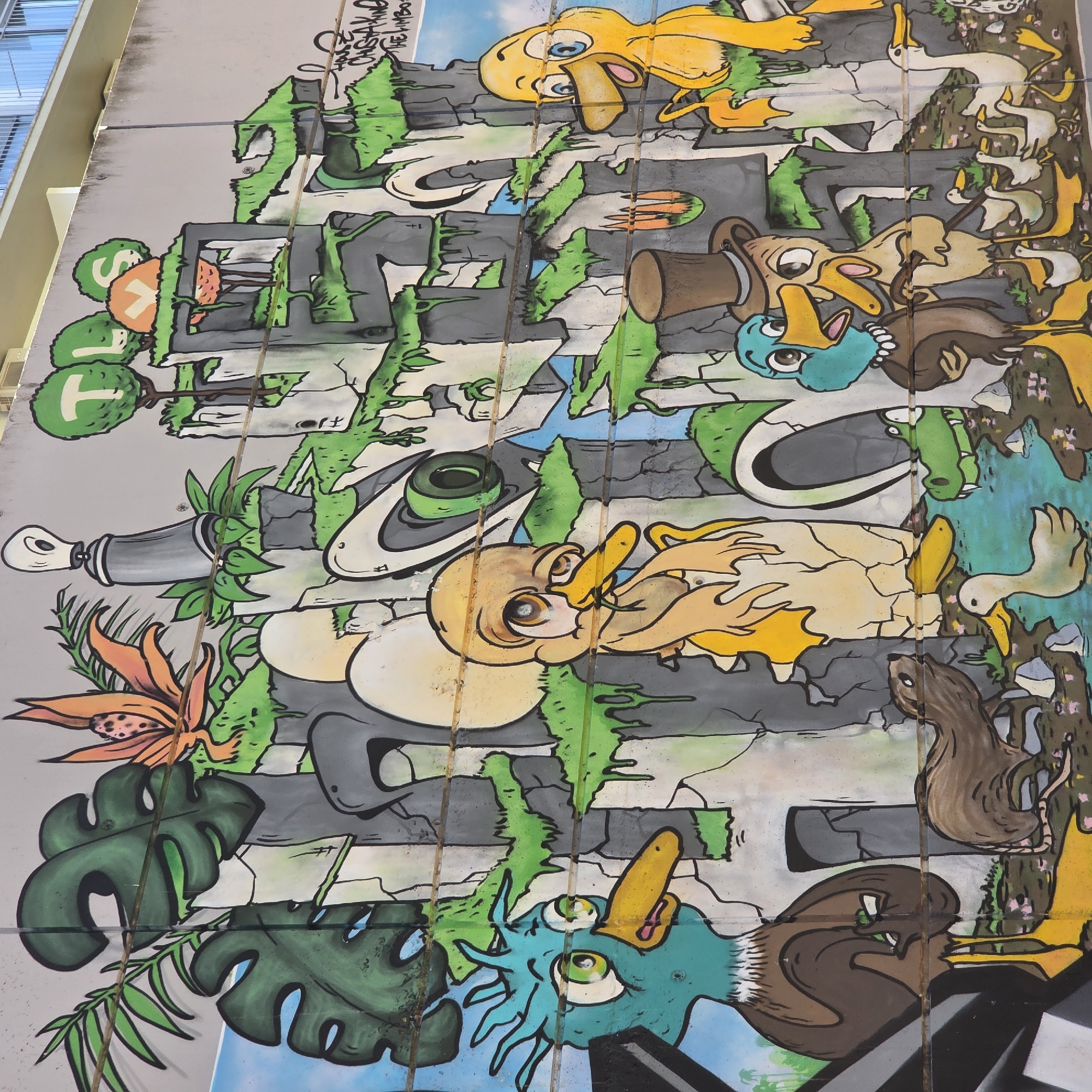 The image shows a section of a large, colorful mural on a building in Guardamar del Segura, Spain. The mural features cartoon-like depictions of ducks in various settings. Some ducks are wearing hats or engaging in human-like activities. The background includes representations of plants, trees, rocks, and water. The artwork is spread across multiple sections of the building's exterior, with lines dividing the panels. The style is vibrant and whimsical, suggesting a playful or educational theme.