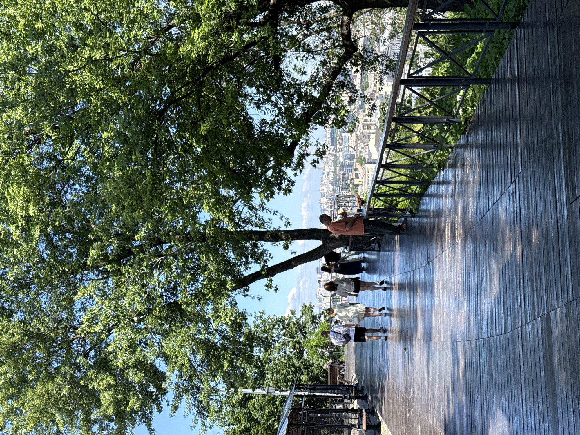 In Kyiv, Ukraine, a paved walkway with a wooden plank surface winds through a park. The walkway is lined with a metal railing and lush green foliage. A large tree with dense foliage dominates the upper left of the frame, casting shadows on the walkway. Several individuals are visible on the walkway. In the foreground, three people walk away from the camera, dressed casually. Further down the walkway, another person stands near the railing, looking out over a cityscape. In the background, a panoramic view of Kyiv's urban landscape unfolds under a clear blue sky with scattered clouds. The overall scene suggests a pleasant day in a public park with scenic views.