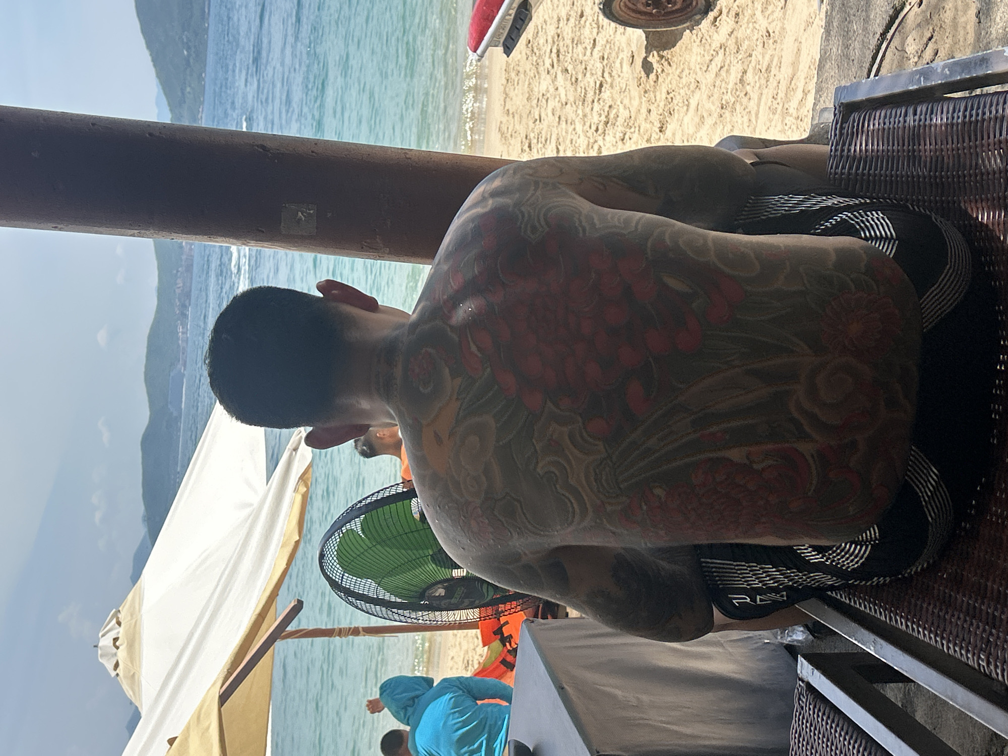 A man with extensive back tattoos sits on a chair facing away from the camera, looking out at the ocean. He is wearing black shorts with white striped accents. To his left, a large green fan is positioned near a table covered with a white cloth. Behind the fan, two people in bright blue and orange clothing are visible on the beach. In the background, the calm blue ocean stretches towards a distant shoreline with green hills and buildings. The scene appears to be a beach resort in Nam Nha Trang Ward, Vietnam, indicated by the umbrellas, beach chairs, and the distinctive landscape. The lighting suggests it is daytime.