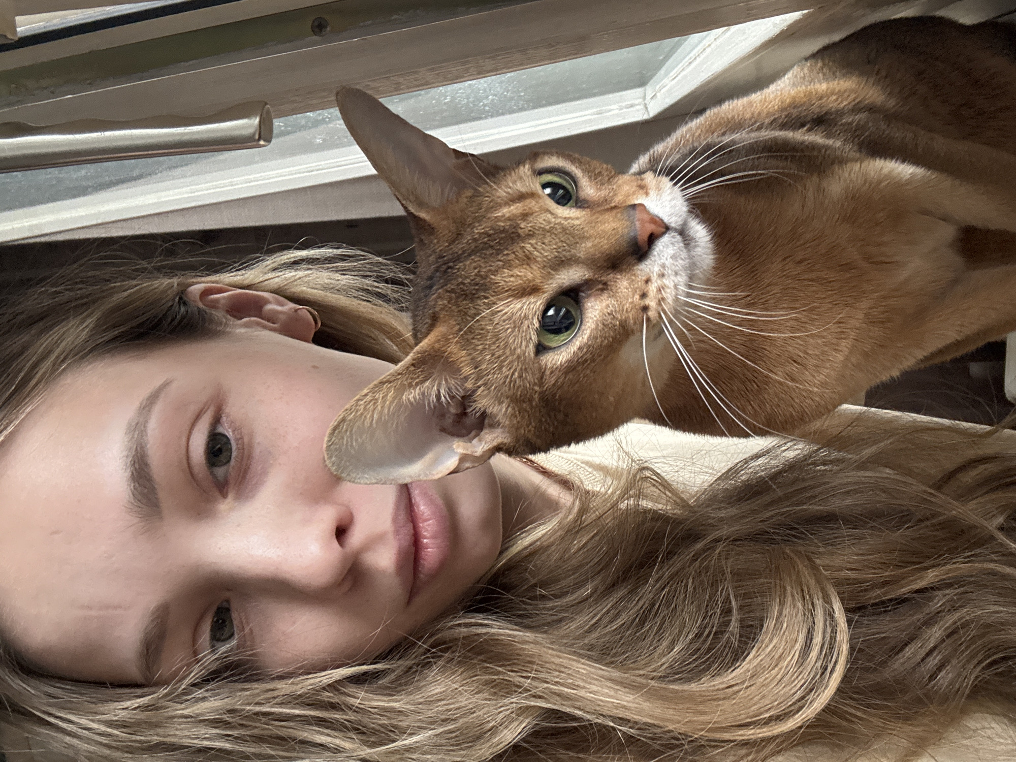 A selfie-style image shows a woman lying on her back with a cat's head positioned close to her face. The woman has fair skin, light brown hair with blonde highlights, and green eyes. Her hair is styled in loose waves and spreads out around her head. The cat, an Abyssinian, has reddish-brown fur, green eyes, and long white whiskers. The cat's ear is close to the woman's face. Above them, a window frame with a metal handle is visible. No information indicates the location is in Chaikovsky, Russia.