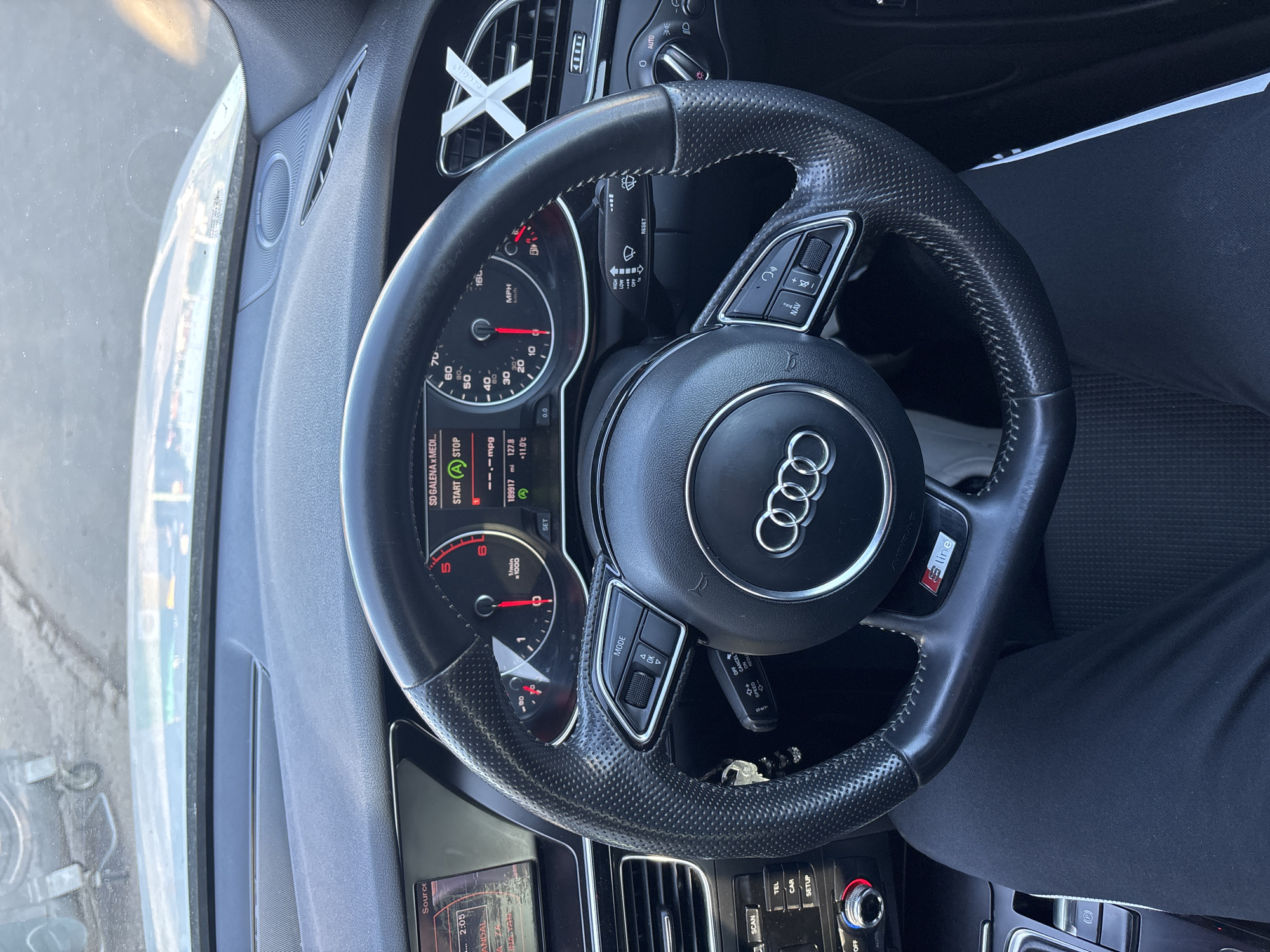 A car's interior is visible, including the steering wheel, dashboard, and center console. The steering wheel has an Audi logo in the center and the S Line badge on one spoke. The instrument cluster shows gauges and a digital display with some information, including "SD GALENA & MEDI" and a speedometer reading. Parts of the center console and dashboard show buttons, vents, and a display screen. A person wearing black pants is seated in the driver's seat, partially visible. The image does not provide enough information to identify the location as Dolni Tsibar, Bulgaria.