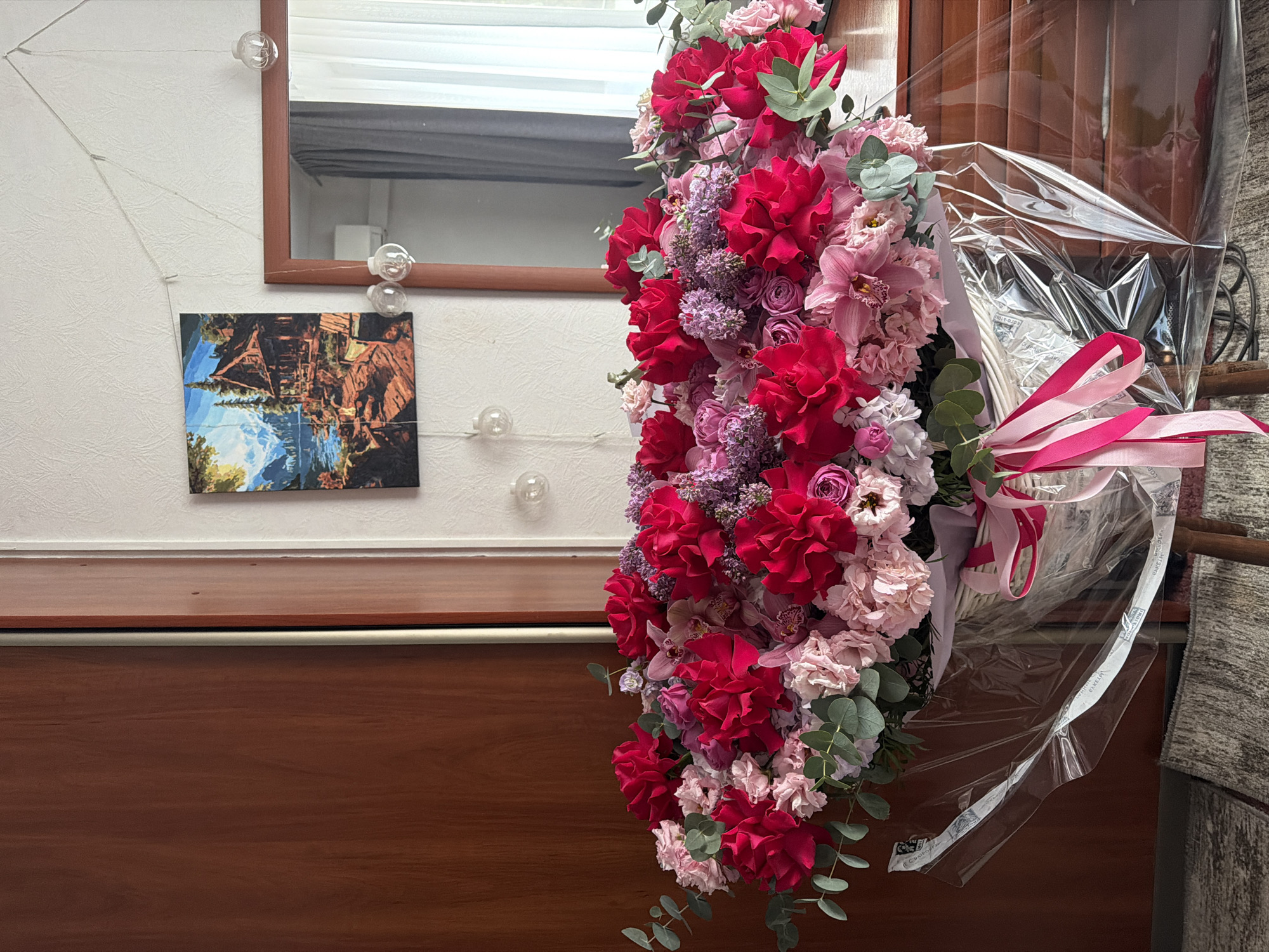 A large floral arrangement, composed of red roses, pink orchids, and light purple flowers, is prominently displayed against a backdrop consisting of a white wall with a landscape painting and a framed mirror, and a dark wood surface. The bouquet is wrapped in clear plastic and tied with a pink ribbon. The overall scene suggests an interior setting, potentially in Moscow, Russia, as indicated by text on the packaging.