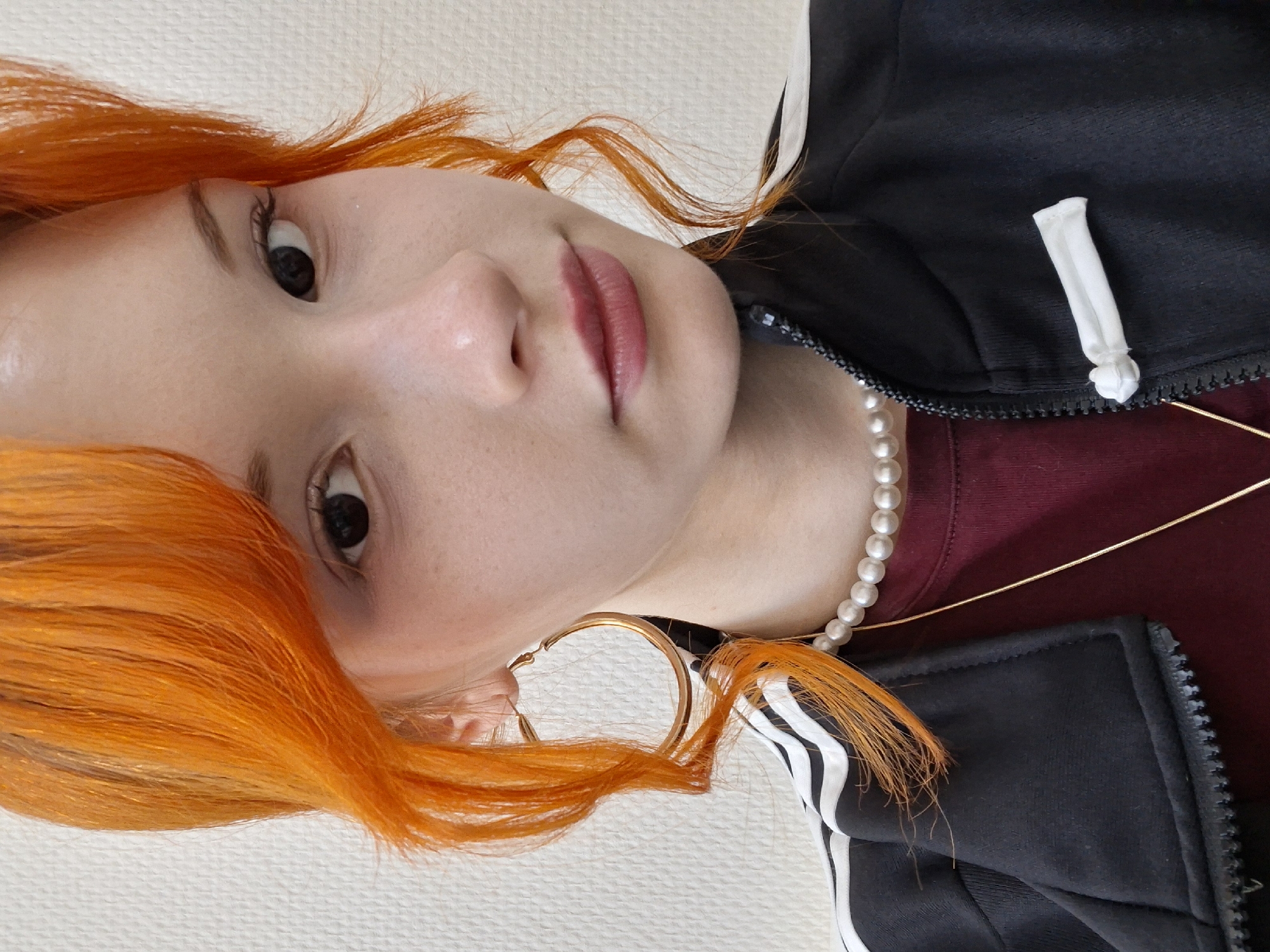 A close-up, rotated view of a person with fair skin and orange hair is presented. The individual has dark eyes and pink lips. The person is wearing a pearl necklace and a large gold hoop earring is visible. They are also wearing a black and white Adidas jacket over a maroon top. The background is a plain white surface. The location is not determinable from the image.