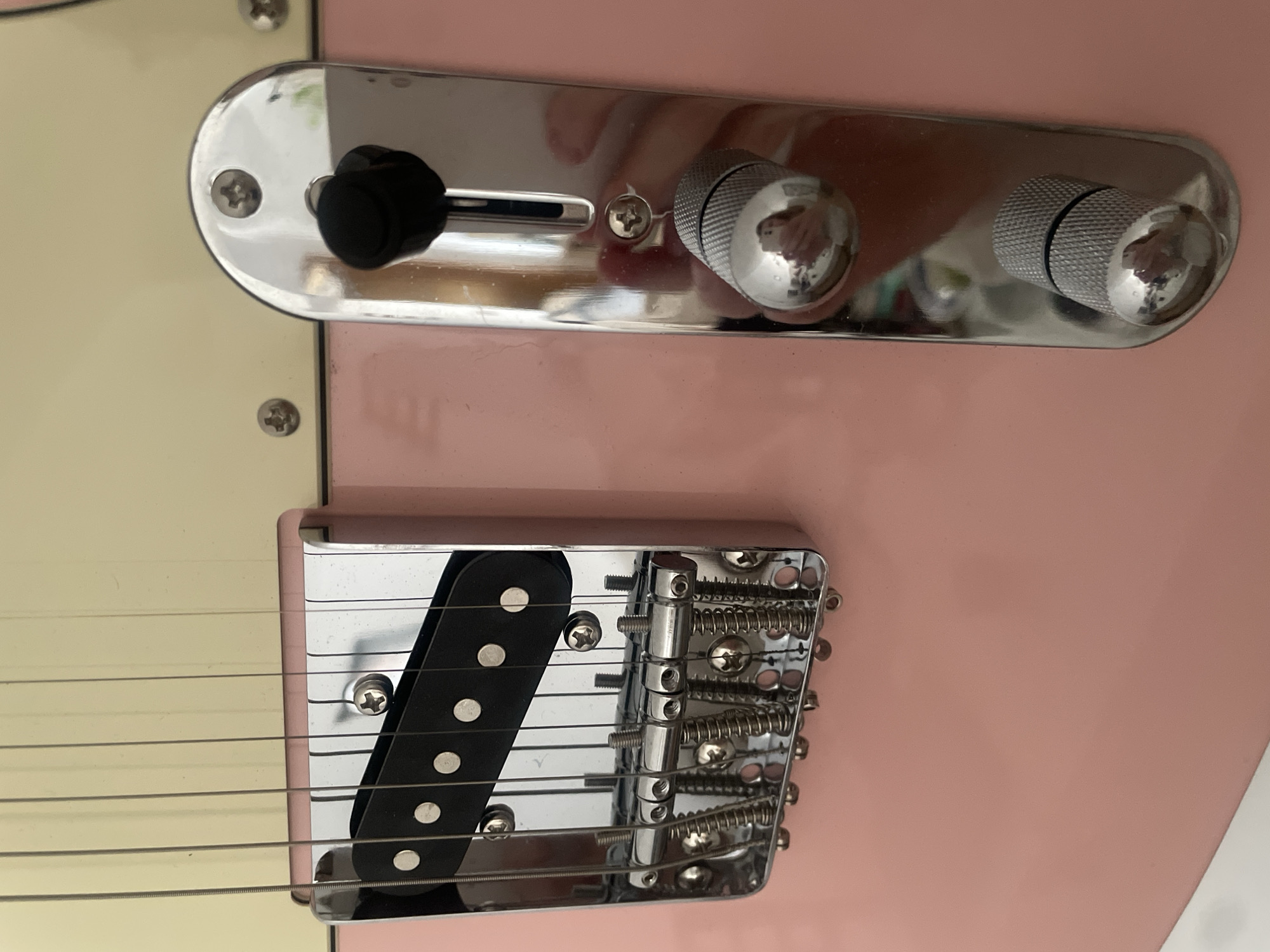 A close-up, eye-level shot shows the hardware of a light pink electric guitar. The guitar has a light yellow pickguard and is lying on a surface that is not fully visible but appears to be white. The guitar's bridge and pickup are in the foreground. The bridge is made of chrome and has six adjustable saddles, each with a small spring. The pickup is black and rectangular, with six silver pole pieces. To the right of the bridge is a chrome control plate with two silver knobs and a black toggle switch. The knobs have a textured surface. The image is well-lit and in focus. There is no indication of the city of Atarfe, Spain, in the image.