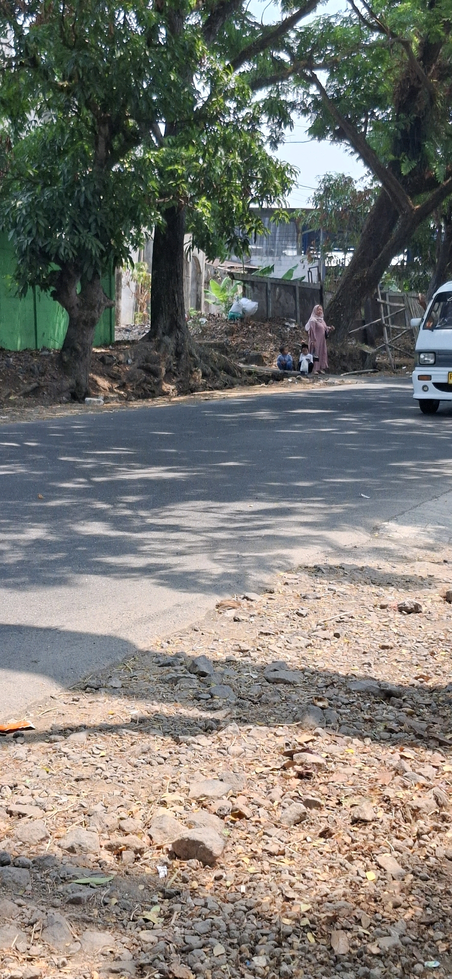 This is a medium shot, eye-level, vertical, outdoor, daytime, natural light, clear day photograph taken on a roadside in Cisalak, Indonesia. In the foreground is a paved road casting many shadows from trees overhead. The ground to the right of the road is covered in rocks and dry leaves. In the middle ground, there are three people sitting near the roots of a large tree. One person is wearing a pink headscarf and a long pink dress. To the left of this person are two younger individuals, one in blue shorts and a white t-shirt, and the other in a white shirt and black pants. Behind them, the ground is uneven and covered with debris. Further in the background, a white vehicle, likely a van, is partially visible on the right side of the frame. The environment suggests a rural or semi-rural setting with lush trees lining the road.