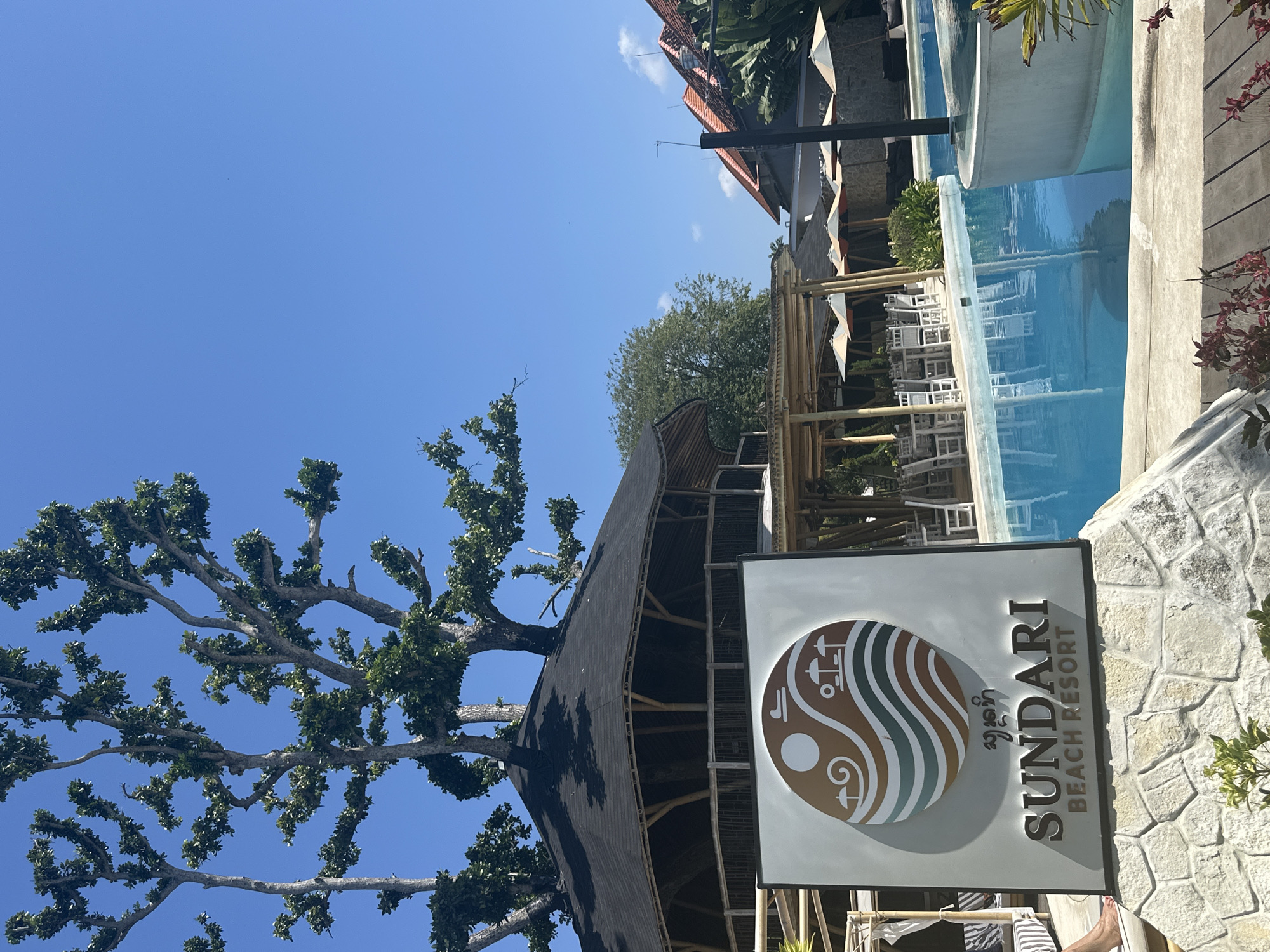 An outdoor shot shows a resort pool area in Denpasar, Indonesia. In the foreground, a sign for "SUNDARI BEACH RESORT" is mounted on a stone base. Behind the sign, a large swimming pool with bright blue water is visible. The pool has an abstract shape with a curved edge. Several white lounge chairs and umbrellas are set up around the pool. In the background, a distinctive circular building with a dark roof and bamboo accents stands under a clear blue sky. Trees with sparse foliage grow out of the roof of this building. Further in the background, more resort buildings with terracotta roofs and lush green foliage are visible. The scene appears to be well-lit, suggesting a sunny day. No people are clearly depicted in the image, though a bare foot is visible on a lounge chair to the left of the sign.
