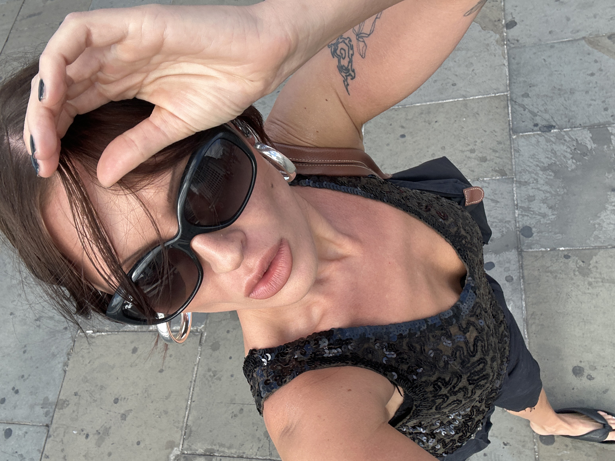 A medium shot, high-angle selfie shows a woman from the chest up, walking outdoors. She has brown hair, sunglasses, and large hoop earrings. She is wearing a black sequined sleeveless top and a black skirt. A brown purse strap is visible over her shoulder. A tattoo is visible on her right arm. Her left hand is raised, touching her hair near her sunglasses. Her right hand is partially visible, and her right foot is visible wearing a black flip-flop. The background is a grey paved surface. The scene appears to be on a street in London, United Kingdom.