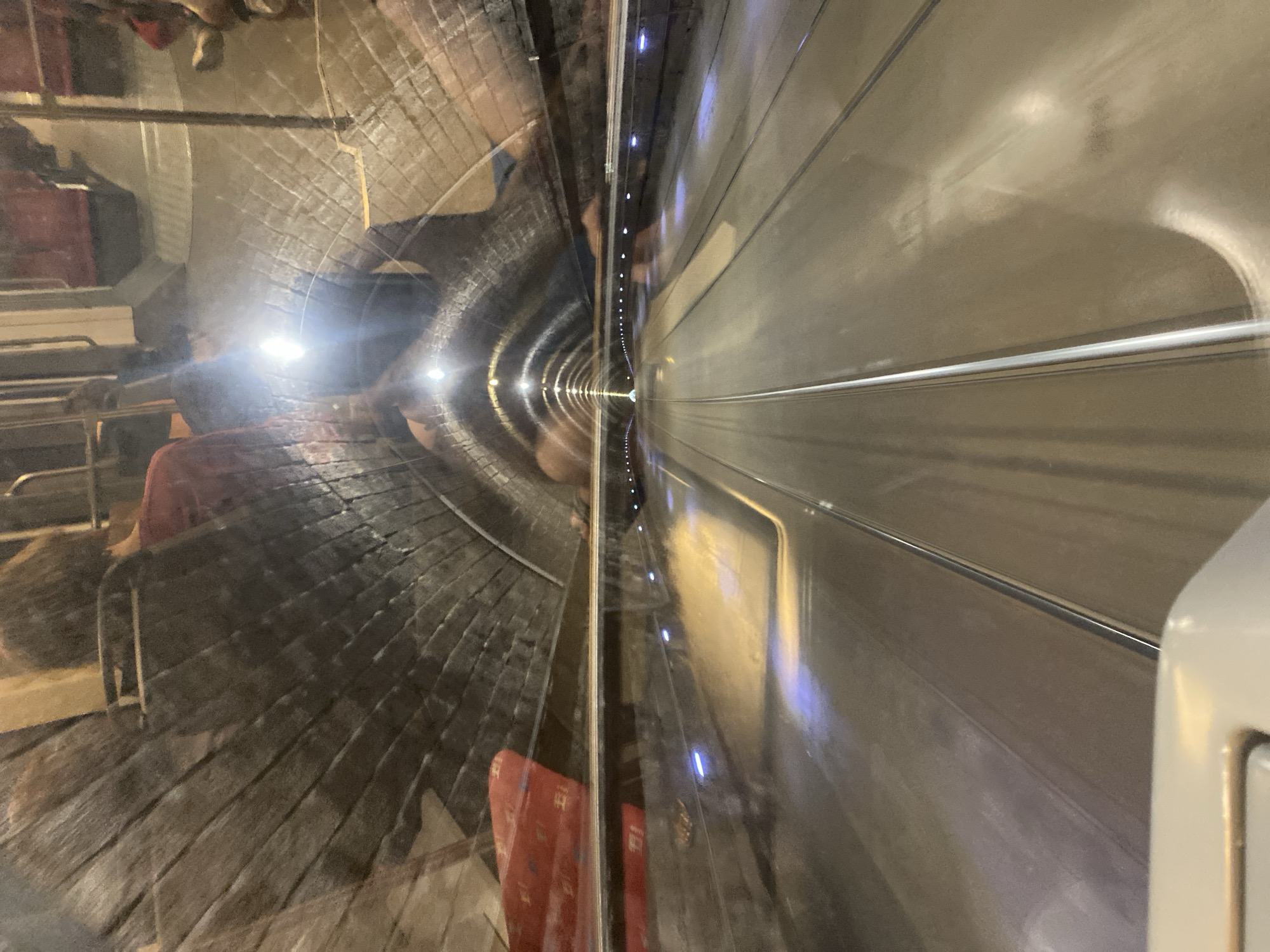 The image captures a distorted, reflective view inside what appears to be a train or subway tunnel. On the left side, a curved tunnel wall constructed of dark bricks is visible, with faint reflections of lights and what seem to be people. The reflections suggest a mirrored surface, possibly the window of the vehicle. The right side of the image shows a smooth, metallic wall of the vehicle's interior, with horizontal lines and a sleek finish. The tunnel recedes into the distance, lined with small, evenly spaced lights that create a sense of depth and perspective. The overall impression is one of motion and transit through an urban underground environment. The lighting is artificial, with bright points from the vehicle's interior and the tunnel lights. Given the location context of Bratislava, Slovakia, it is likely this is a subway tunnel within the city. There is no visible text in the image.