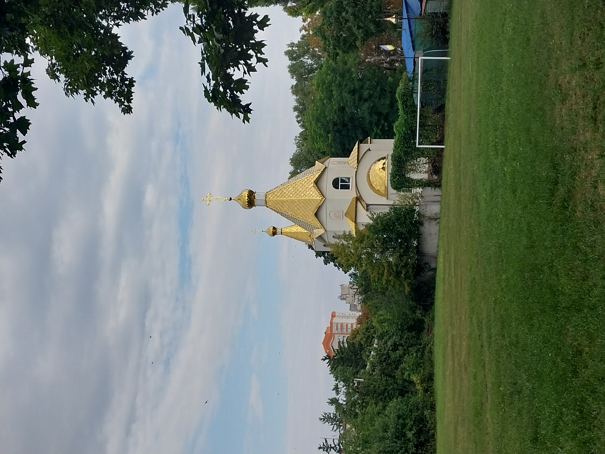 In the city of Doslidne, Ukraine, a serene scene unfolds with a domed church as the focal point, situated amidst lush greenery. The church, adorned with golden domes and crosses, stands out against a backdrop of trees and a partly cloudy sky. In the foreground, a grassy field with a goalpost suggests recreational activity. The overall atmosphere is peaceful, with hints of urban life in the form of distant buildings. The image captures a tranquil moment in a Ukrainian town, blending religious architecture with natural beauty and everyday elements.