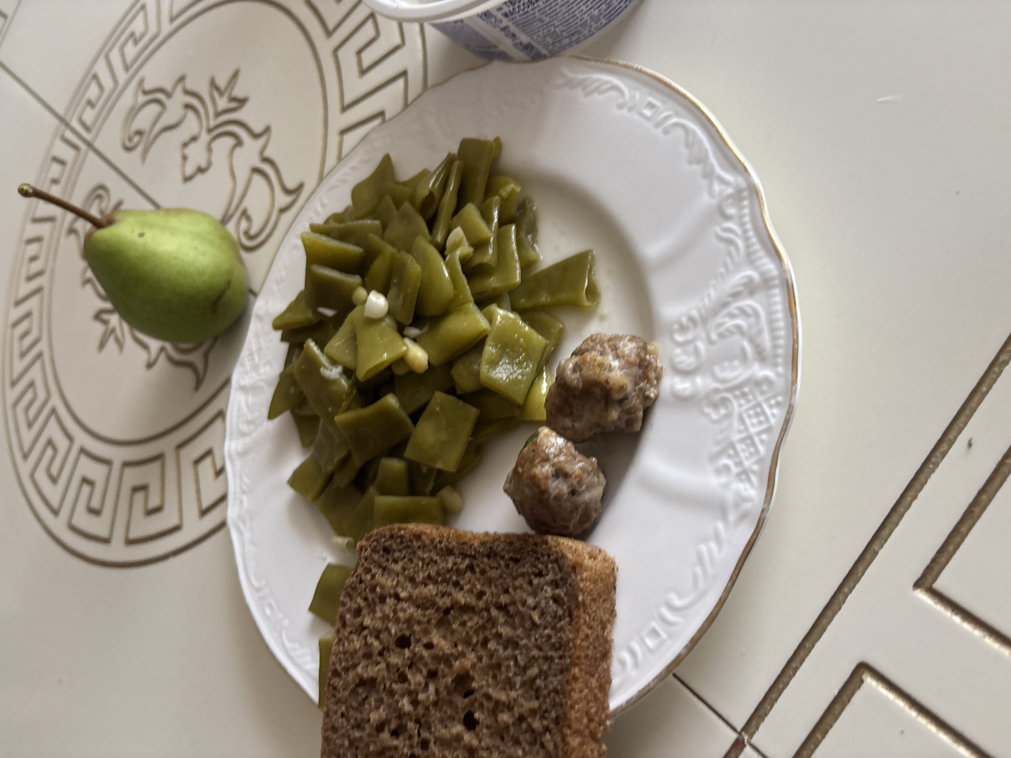 A white plate with a gold rim sits on a light-colored, tiled surface. The plate contains a serving of cooked green beans, two small meatballs, and a slice of dark bread. To the upper right of the plate, a single green pear rests on a larger decorative plate or placemat with a Greek key pattern. The tiled surface has a geometric design in gold. A partially visible blue and white patterned container is on the right edge of the image. The scene appears to be a meal setting.