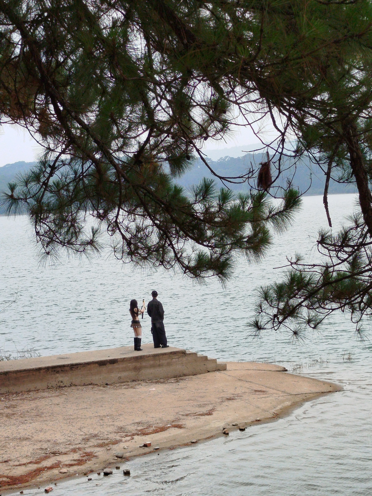 A medium shot shows two individuals standing on a concrete pier that juts into a body of water, likely a lake, in what appears to be Da Lat, Vietnam. A woman wearing a short skirt and knee-high boots stands facing the water, holding an object that is difficult to discern. A man wearing dark clothing stands beside her, also facing the water. The background consists of the lake, with distant hills visible on the horizon. The scene is framed by a pine tree branch that hangs down from the top of the frame, partially obscuring the view. The weather appears to be overcast. The pier is made of concrete and is located on a sandy beach. There are bricks and rocks scattered on the ground near the water's edge. The water appears to be calm.
