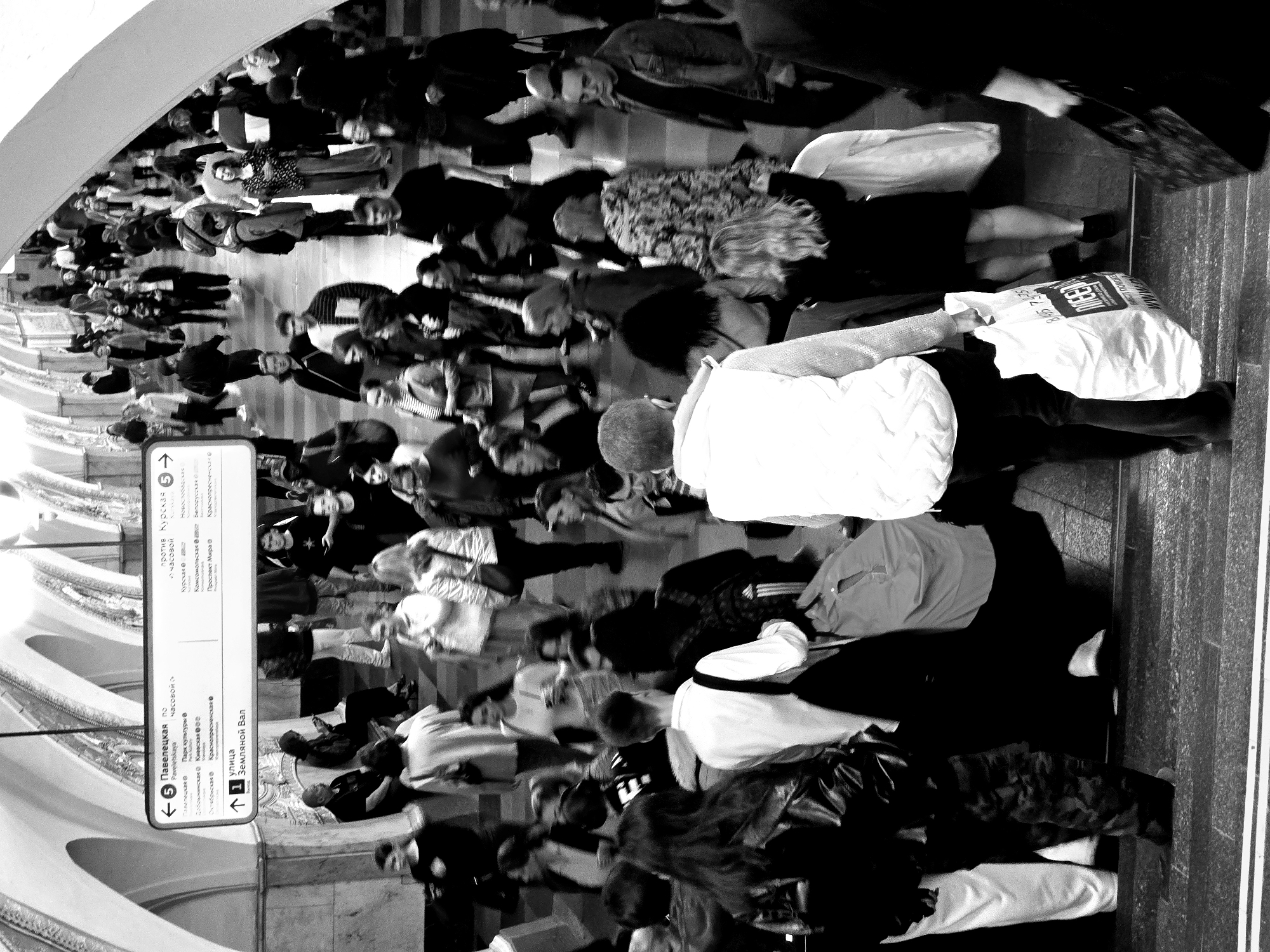 The image is a black and white photograph taken in what appears to be a subway station in an unknown city. The composition features a crowd of people walking, suggesting a busy, public transportation environment. The presence of a directional sign provides information about the station's connections, with text in a language other than English, giving clues about the location. The sign indicates destinations like "Павелецкая" and "Курская" along with other station names. The crowd consists of people of varying ages and outfits. The photograph also includes an advertising banner or sign with visible text, with recognizable English text with "ВМЕСТЕ" and numbers "8,445 74-75". The environment includes architectural details such as arched ceilings.