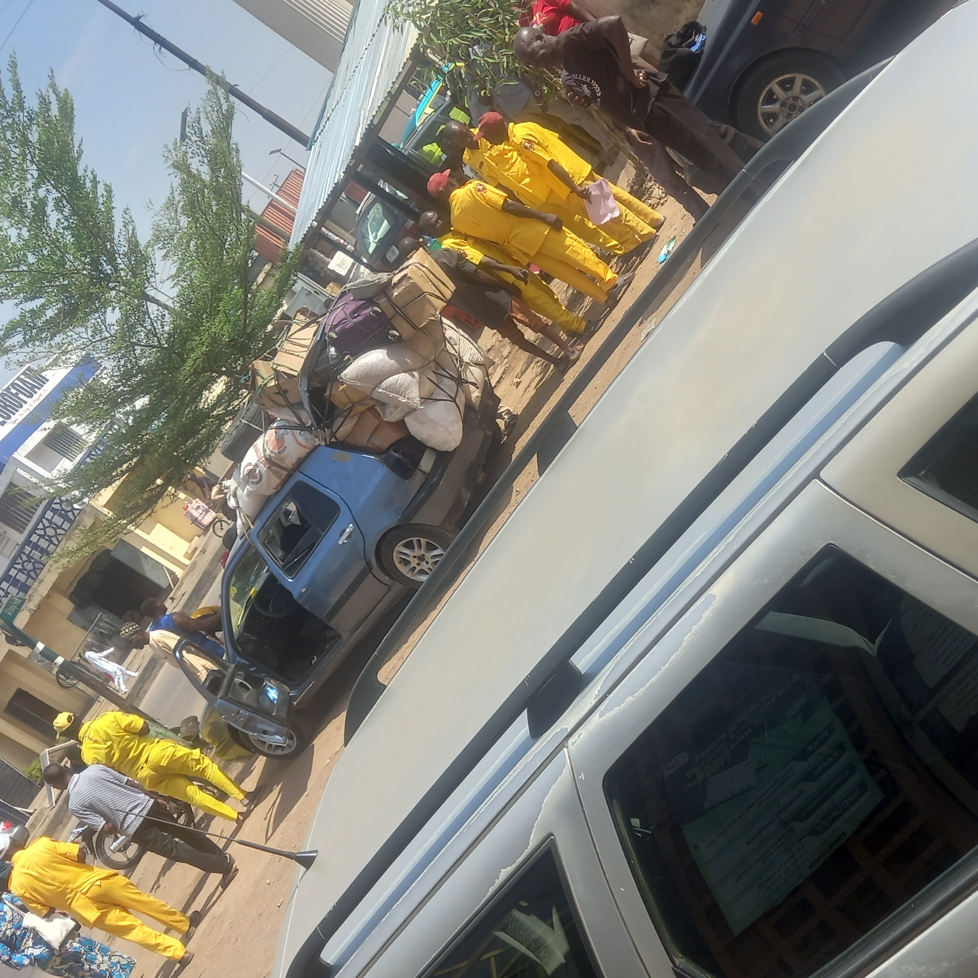 This image captures a bustling outdoor scene in Dadi Riba, Nigeria, under a bright, clear daytime sky. The perspective is slightly elevated and tilted, looking down at a street or open area filled with people and vehicles.
In the immediate foreground, the side of a light grey van or SUV is visible, featuring a roof rack. Through its window, a document or poster with text is partially discernible, showing words like "VISA," "COMMUNITY," "PROGRAMME," "9000," and "ACC."
Further back and to the left, a blue sedan or hatchback is prominently featured, heavily overloaded with various items, including large white sacks and cardboard boxes, meticulously tied to its roof. Its driver's side door is open. Surrounding this vehicle are numerous individuals. A significant group of people are seen wearing distinctive bright yellow uniforms, some complemented by red caps, suggesting they might be part of an organized group, a workforce, or officials. One person in a striped shirt stands near the blue car, holding a long, dark pole. Other individuals are dressed in casual clothes, including one man with a black t-shirt bearing the word "MILLER."
The background features green trees and various buildings. One prominent building clearly displays the text "EUROFOAM" in large blue letters. Elements of corrugated metal roofing are also visible on some structures. The ground appears to be unpaved in parts, adding to the urban, possibly market-like atmosphere. The overall scene depicts a lively and active street environment, characteristic of a busy day.