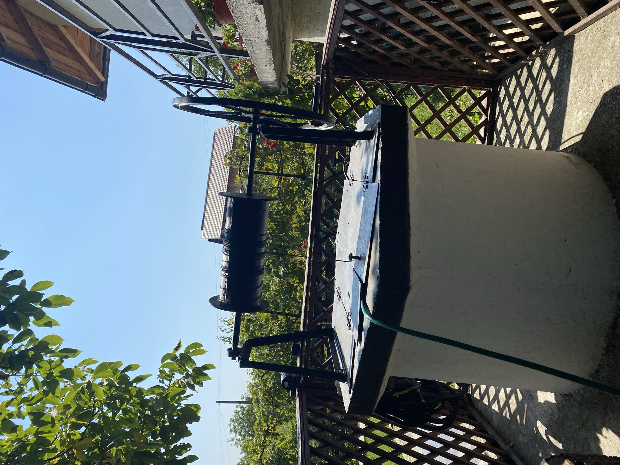 The image depicts an outdoor scene featuring a well, a building with a balcony, and lush greenery under a clear blue sky. The well, made of white concrete with a black rim, is centrally located. It has a metal frame supporting a black winding mechanism, likely for drawing water, with a large spool and a hand crank. A green hose snakes from the top of the well and lies on the ground. The well is situated on a concrete patio surrounded by a brown wooden lattice fence.
To the right, a building's concrete balcony with a black metal railing is visible, adorned with potted red flowers. Part of the building's roof, made of wooden shingles, is also in frame. In the background, trees and foliage create a dense green backdrop. The overall impression is a rustic, residential setting.
While the image provides a detailed view of the well and its surroundings, there is no information within the visual data to ascertain its specific location within the city of Târgu Neamț, Romania. The scene appears to be a private property. No people are present in the image, and no actions or interactions are depicted.