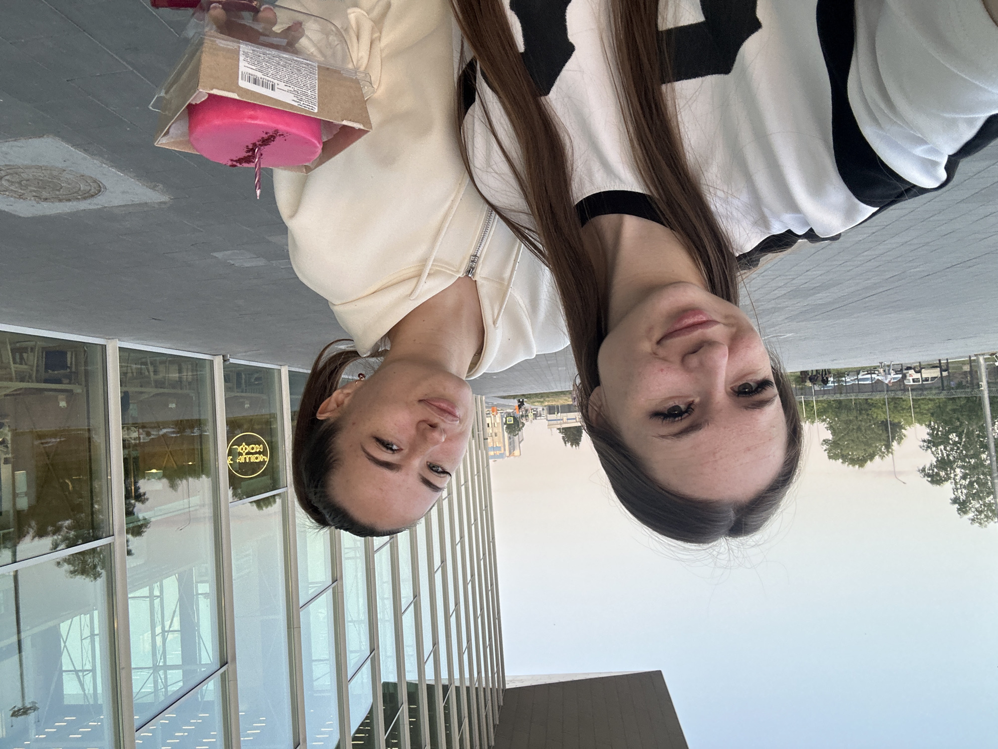 Two young women are posing for a selfie outdoors in front of a modern building with a glass facade. The woman in the foreground on the left is wearing a black and white long-sleeved shirt and has long brown hair. The woman behind her on the right is wearing a cream-colored zip-up hoodie and has her hair in a ponytail. She is holding a small, round, bright pink cake with a single lit candle in a cardboard container. The background shows a paved area, some trees, cars, and the exterior of the building which reflects the sky and surrounding environment. The sky is a pale blue, indicating it is likely late afternoon or early evening.
