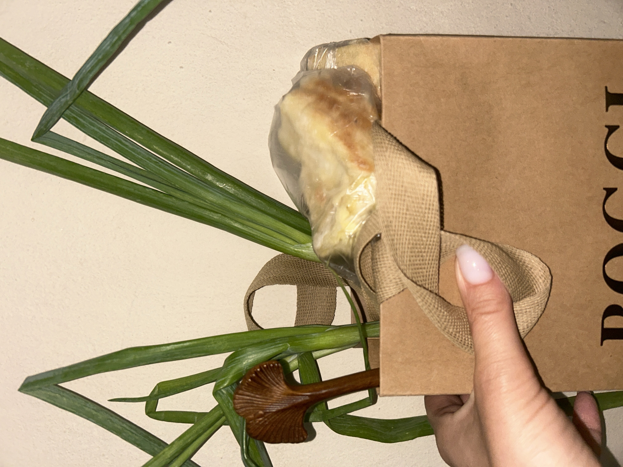 In Buhovo, Bulgaria, a light-skinned human hand, with a manicured thumbnail painted a pale pink, grips the upper left edge of a light brown kraft paper bag. The bag features a woven beige fabric handle, part of which is secured by the thumb. Visible dark brown letters at the bottom of the bag spell "POGGI". Protruding vertically from the bag's opening are multiple long, green stalks, characteristic of spring onions or chives. Partially nestled among these stalks is a dark brown, wooden implement with a fan-shaped, ribbed head, resembling a ginkgo leaf or mushroom, and a slender handle. To the right, within the bag, a pale yellow, irregular-shaped food item, possibly a baked good, is encased in transparent plastic wrap. The background is a textured, light off-white wall. Illumination appears to originate from the left, creating minor shadows.
