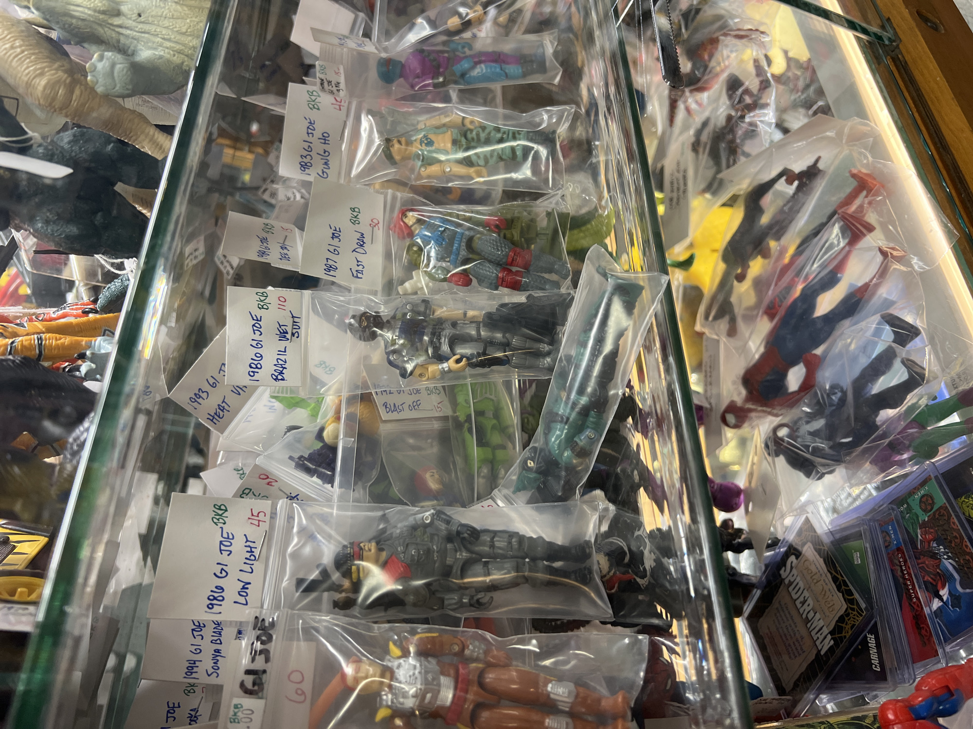 This is a photograph taken within a shop, likely an antique or collectible store, in the City of Newburgh, United States of America. The image captures a display case containing various vintage action figures and trading cards. The figures, individually packaged in clear plastic bags, are primarily G.I. Joe characters from the 1980s and 1990s, as evidenced by the handwritten tags labeling the characters and their respective years (e.g., "1986 GI Joe Low Light," "1983 GI Joe Gung Ho"). The tags also include prices. The bottom right corner of the display case includes several Spider-Man trading cards. The case is made of glass, resulting in reflections. Other items such as dinosaur figures are seen behind the glass of the case. The lighting suggests an indoor environment.