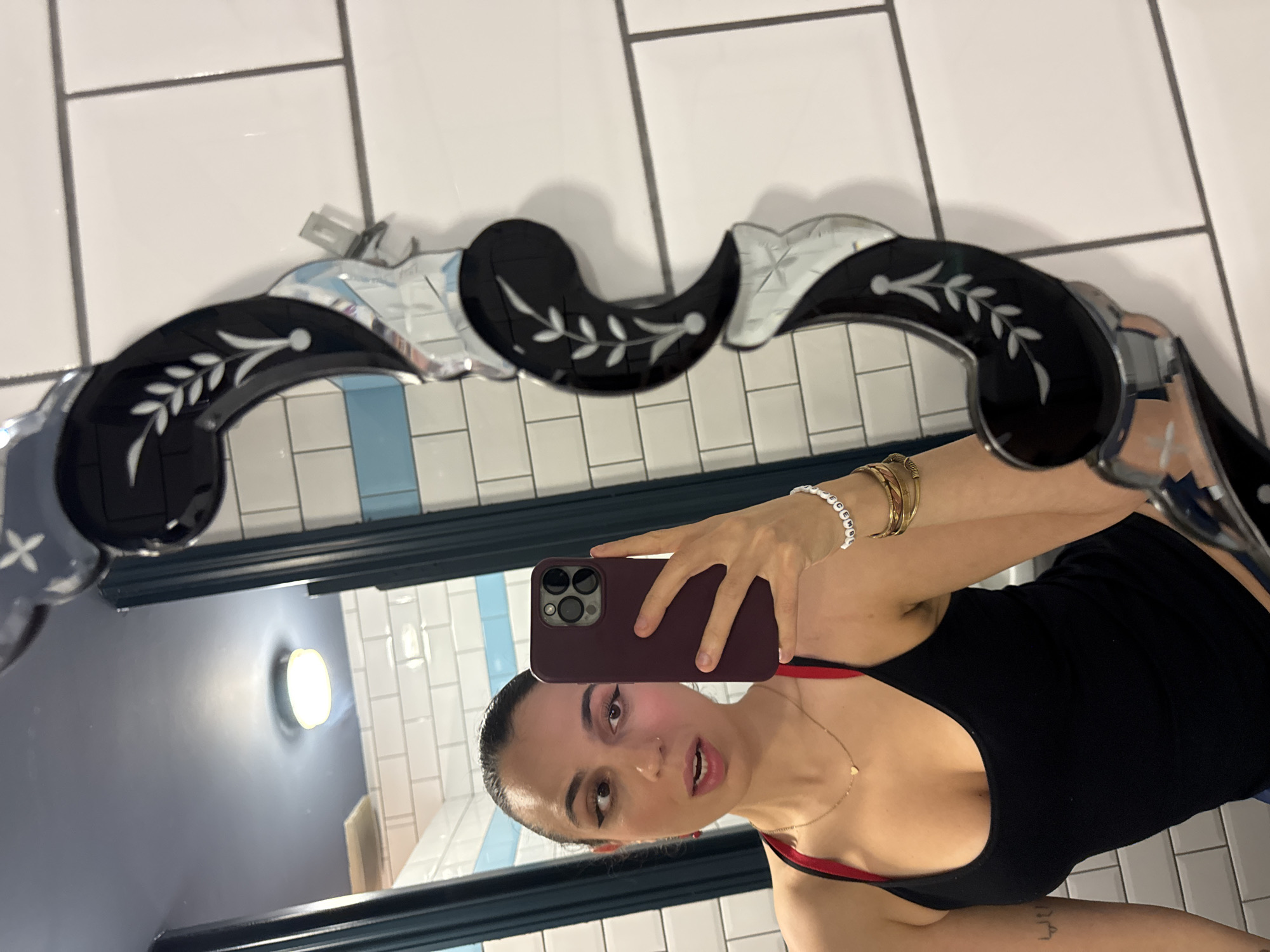 A woman is taking a selfie in a bathroom mirror. The bathroom has white tiled walls and a blue trim. The mirror has a decorative black and mirrored frame. The woman is wearing a black top with red straps and several bracelets. She is holding a burgundy-colored smartphone in her right hand. The mirror reflects a round light fixture on the wall and more white tiles. Based on the style of the bathroom and mirror, the location may be in a historical or tourist district within the city of London, United Kingdom.