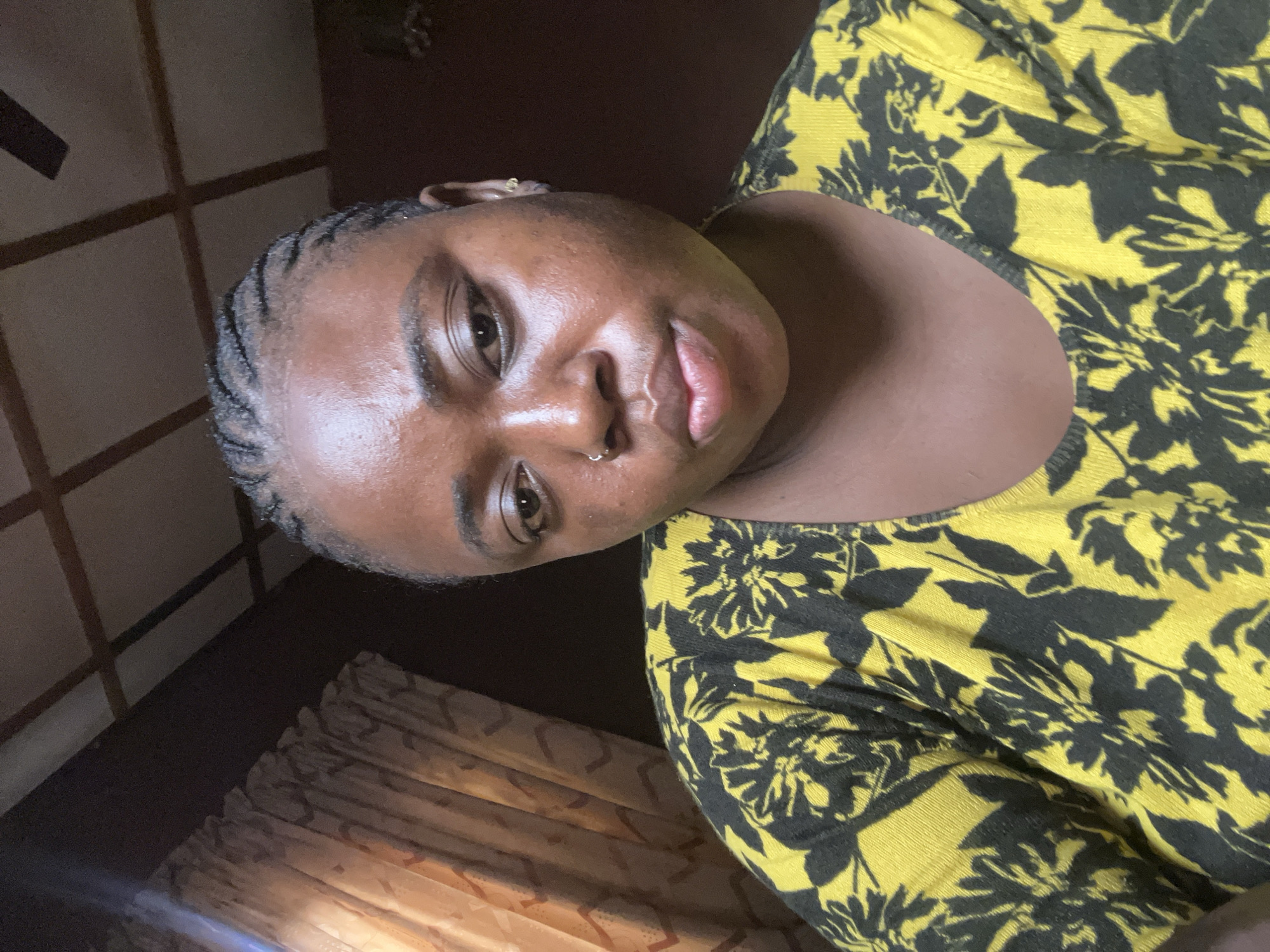 A close-up portrait shot shows a Black woman indoors. Her hair is styled in cornrows. She wears a yellow and black patterned top. A nose ring is visible in her left nostril. The background includes a dark brown wall with a lattice design and a window covered by brown curtains. The setting appears to be a room, possibly in a residential area. Information about the location of Jos, Nigeria cannot be derived from the image.