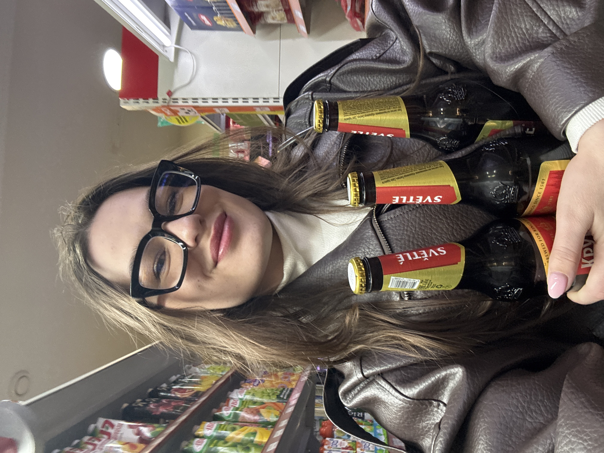 The image shows a young woman wearing glasses and a brown leather jacket, holding three bottles of Czech beer called "Krusovice Světle" (Light Krusovice). She is standing in a store, as evidenced by the shelves of packaged goods visible in the background. The Cyrillic script on the beer labels suggests this image was taken in a location that uses the Cyrillic alphabet, which includes Moscow, Russia.