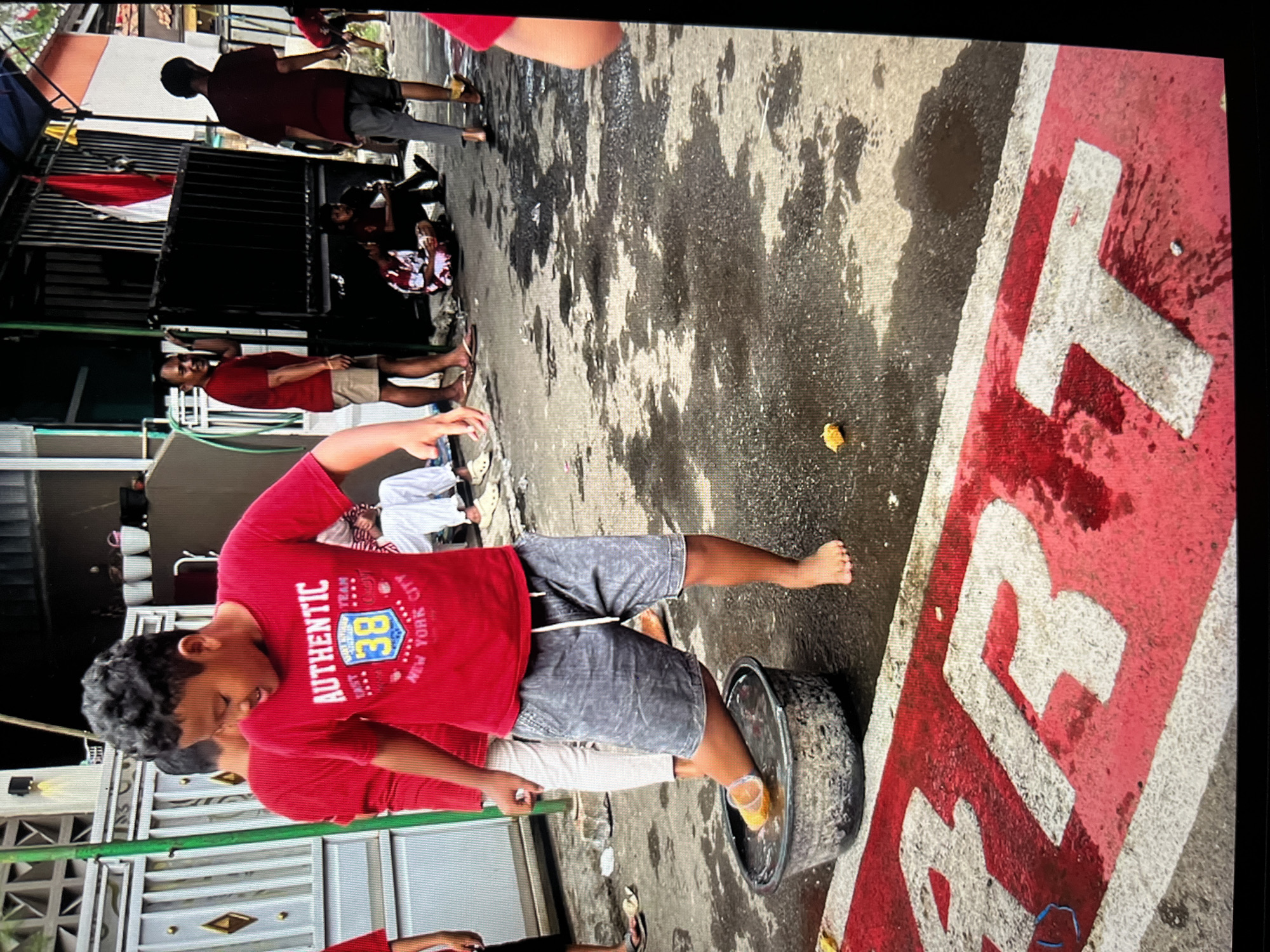 The image depicts a group of people in what appears to be an outdoor urban setting, possibly in the city of Cirahayu, Indonesia, indicated by the presence of Indonesian flags. In the foreground, a young boy wearing a red t-shirt with the text "AUTHENTIC 38 NEW YORK CITY" and grey shorts is standing with his left foot in a large, shallow basin filled with water. He is barefoot except for what appears to be a yellow object on his foot. The ground is wet and appears to be asphalt, with red and white painted markings, possibly denoting a road or pathway. Several other individuals are visible in the background, some standing and some sitting. One man in a red t-shirt and shorts is leaning against a black metal gate, and another person in a red t-shirt and grey pants is standing nearby. Other figures are partially visible, suggesting a gathering or activity. The environment includes buildings and structures, with some vegetation visible. The overall atmosphere suggests a casual, possibly festive or communal event.
