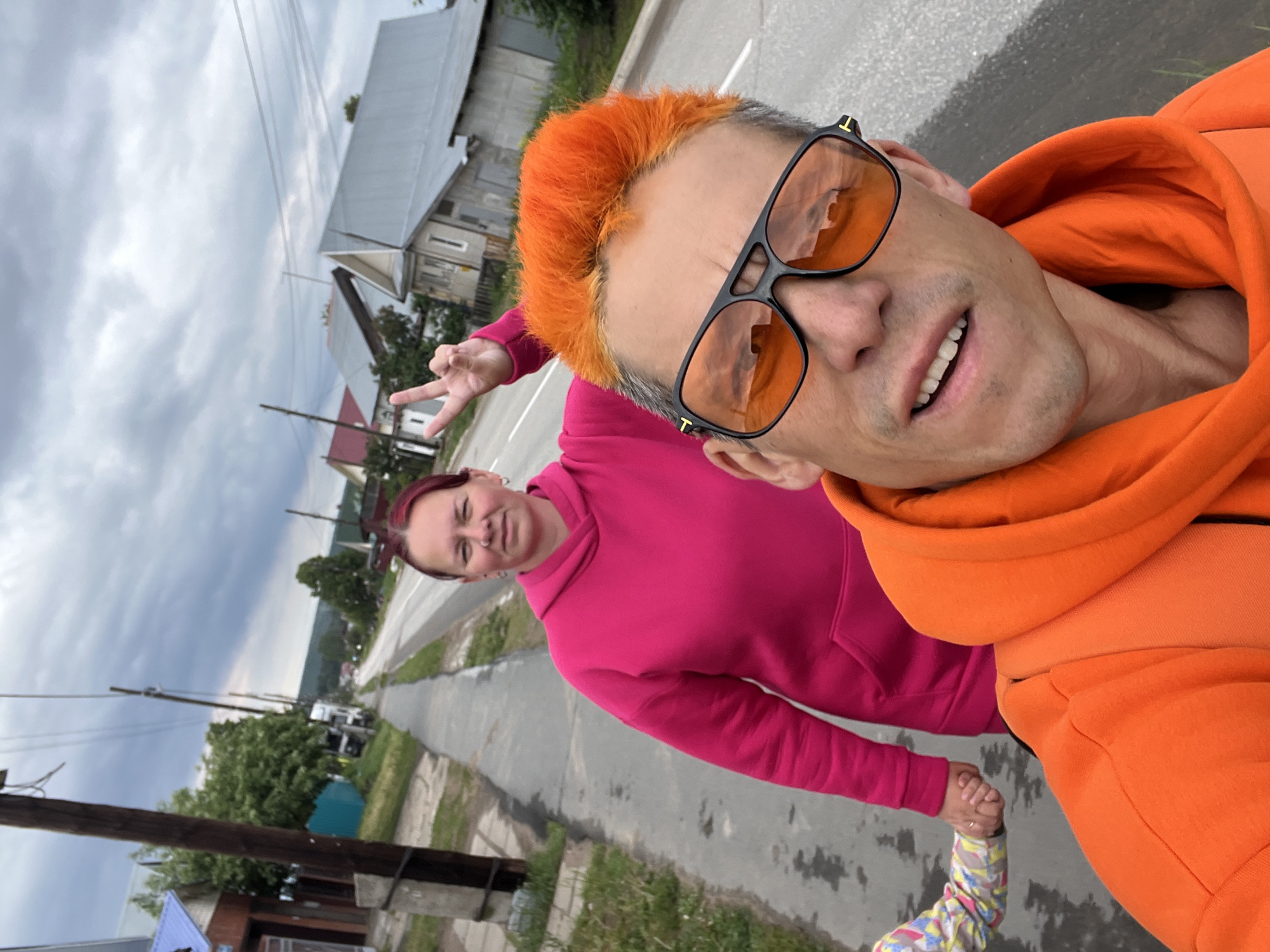 The image shows a selfie taken by a man with dyed orange hair and orange sunglasses, wearing an orange hoodie. He is holding hands with a child who is partially visible at the bottom of the frame. Behind him is a woman wearing a pink hoodie, making a peace sign with her hand. They are walking on a paved road. In the background are houses and trees. The sky is overcast. Based on the available information, it is impossible to pinpoint the exact location within the city of Chaikovsky, Russia.