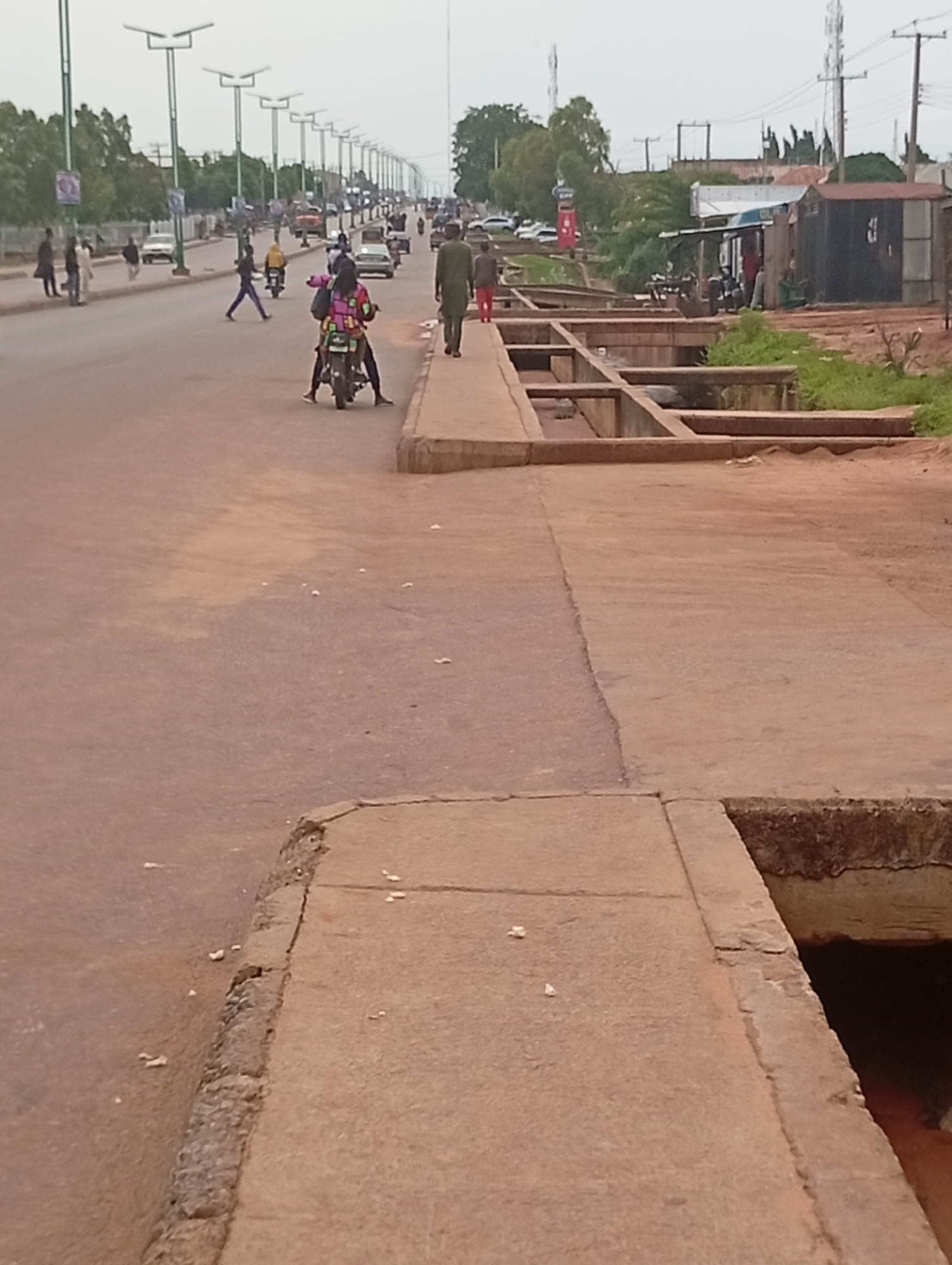 The image depicts a street scene in Tumfure, Nigeria, characterized by a long, straight road lined with streetlights on both sides. The road is paved and appears to be moderately busy with vehicular and pedestrian traffic. Several cars are visible on the road, along with motorcycles and people walking.
In the foreground, to the right of the image, there is a concrete sidewalk or raised pathway adjacent to a drainage channel. The channel is partially visible and contains some reddish-brown water or mud. The pathway itself is made of concrete slabs and shows signs of wear and tear, with some crumbling edges.
Further down the road, there are various structures, including what appears to be small shops or market stalls on the right side of the image. These structures have corrugated metal roofs and some have awnings. Trees and vegetation are present on the sides of the road and behind the buildings, indicating a semi-urban or suburban environment.
The overall lighting suggests it is daytime, possibly overcast, as there are no strong shadows. The image captures a slice of everyday life in this Nigerian city, highlighting its infrastructure, transportation, and commercial activities.