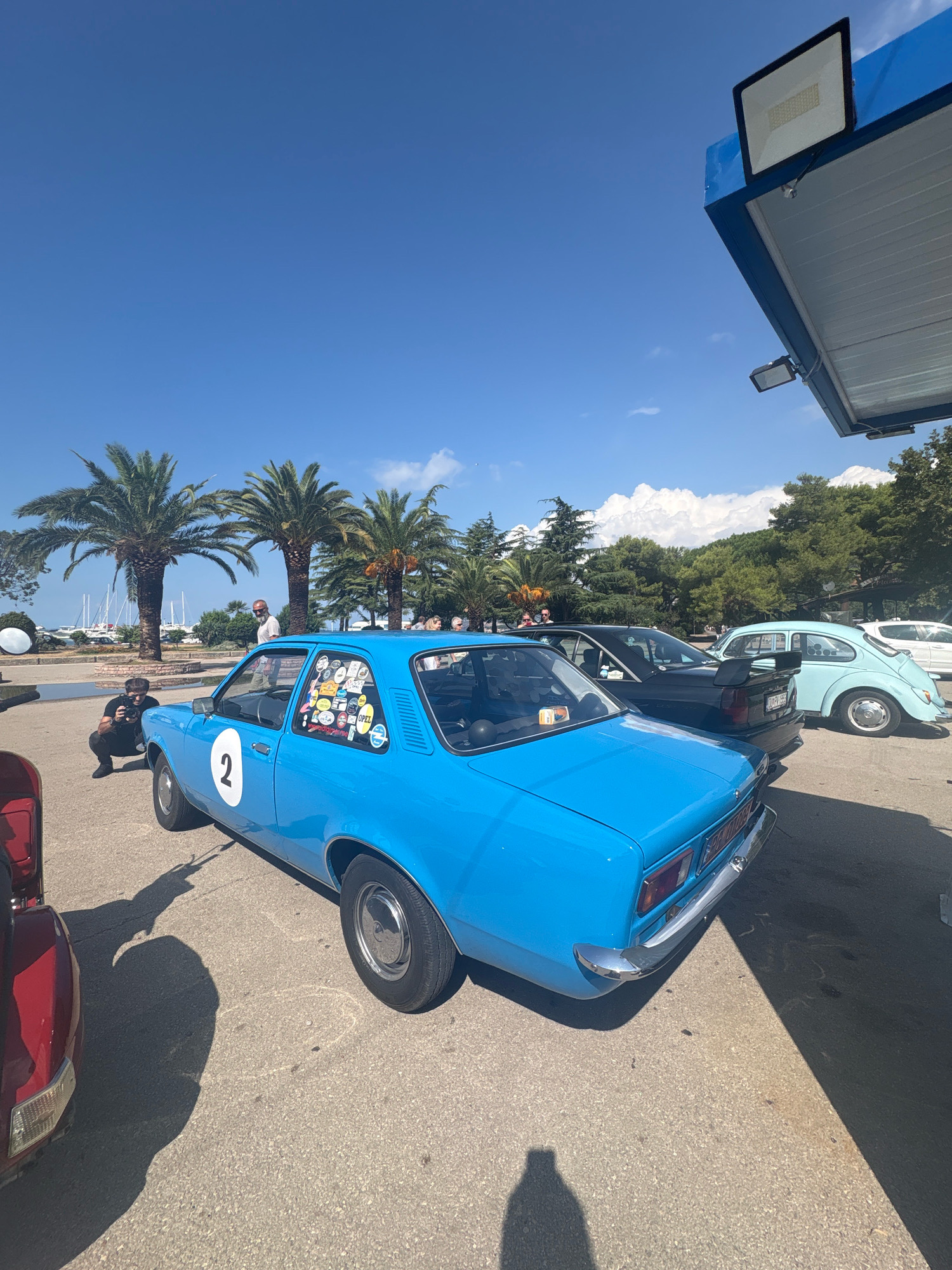 A bright blue vintage Opel Kadett is parked on an asphalt surface, adorned with numerous stickers on its rear window and side. A large white number '2' is visible on the driver's side door. A person is crouching in front of the car, holding a camera and appearing to take a photo. In the background, a man stands near a black car with a spoiler, and further back, a light blue Volkswagen Beetle is parked. Palm trees and other greenery line the area, with sailboats visible in the harbor beyond. The scene is set under a clear blue sky with scattered white clouds, possibly indicating a sunny day in the city of Bar, Montenegro. The cars are parked in what appears to be an open area, possibly a parking lot or a promenade, with a structure providing shade overhead.