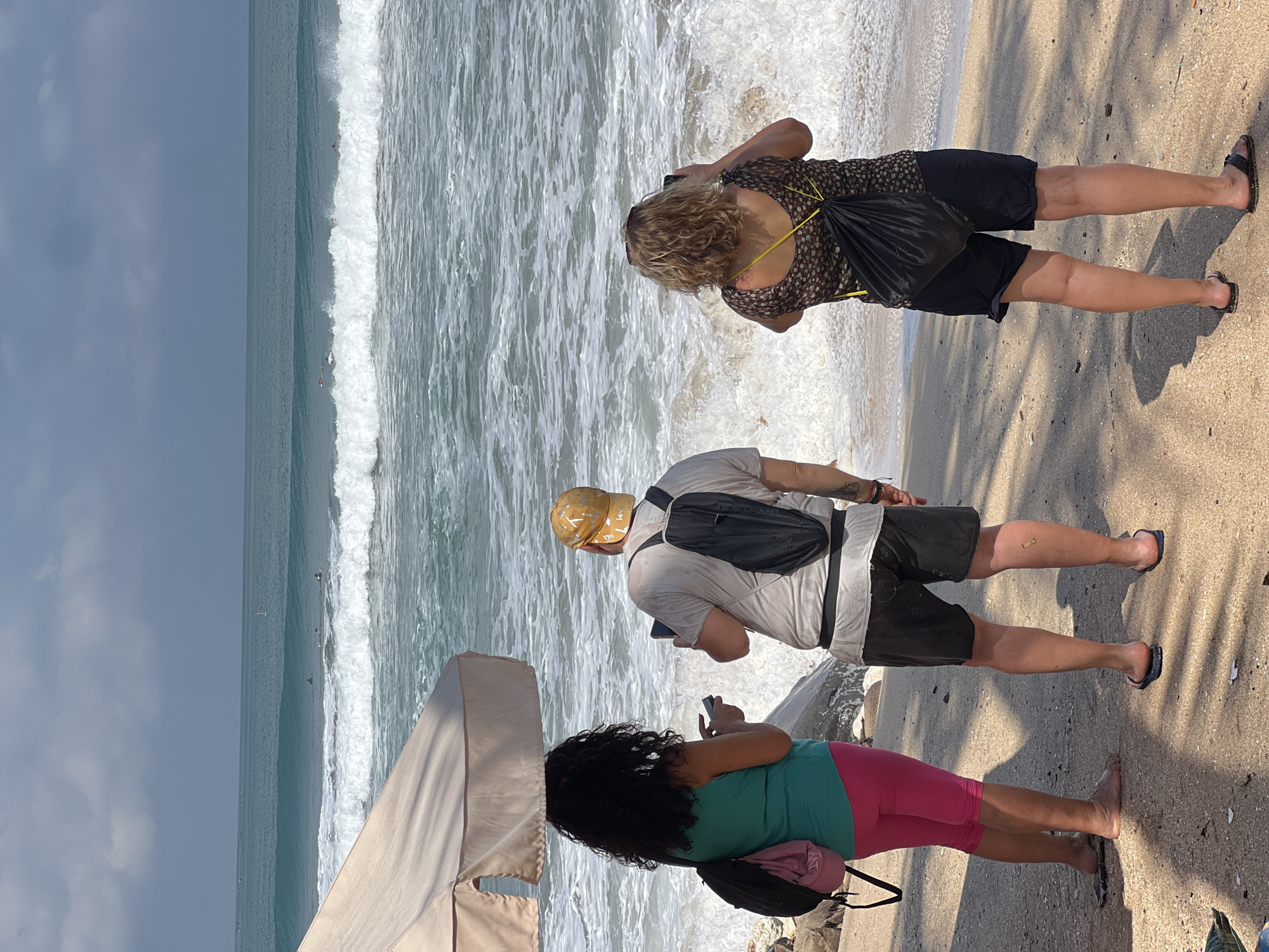 Three people stand on a sandy beach in Kuta, Indonesia, facing the ocean. The person on the left, with curly dark hair and wearing a teal tank top and pink capri pants, holds a phone in their left hand. In the center, a person wearing a light-colored t-shirt, black shorts, and a yellow baseball cap holds a tablet in their right hand. To the right, a person with blonde, curly hair, dressed in a patterned top and black shorts, holds a phone up to take a picture. All three are wearing backpacks. The ocean has moderate waves with surfers visible in the distance. A partially visible umbrella provides shade for the two people on the left. The sand is light brown, with some scattered shells and shadows from palm fronds. The sky is partly cloudy with a mix of gray and blue.